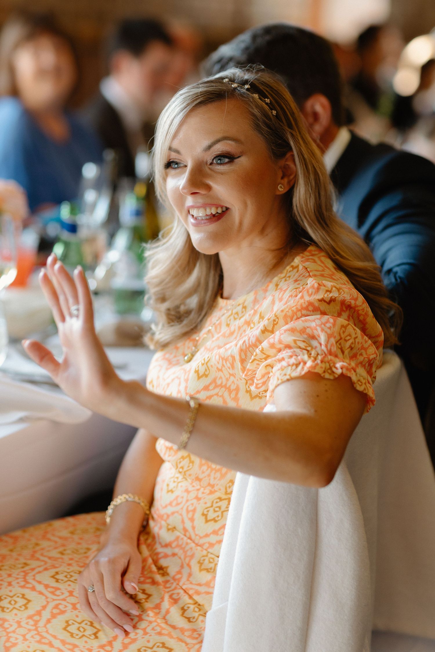 Smiling blonde wedding guest in orange floral dress waves at reception dinner table.