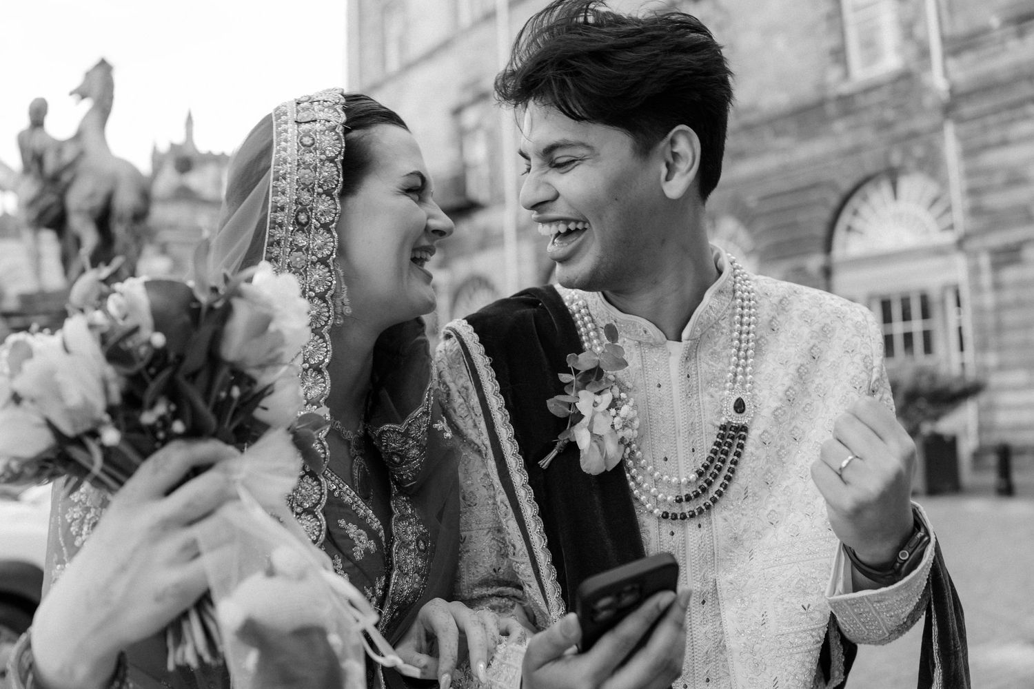 Couple in traditional South Asian wedding attire laughing together outdoors in black and white photo.