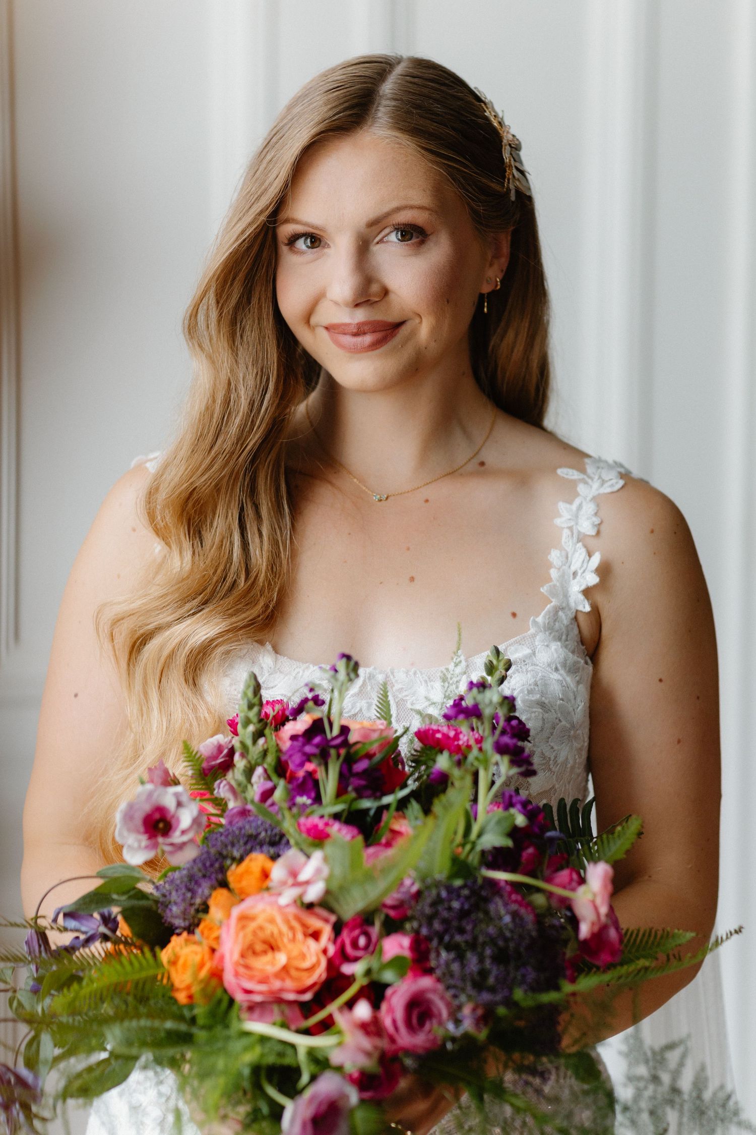 Bride in white lace dress holding colorful purple and orange wedding bouquet, smiling softly indoors.