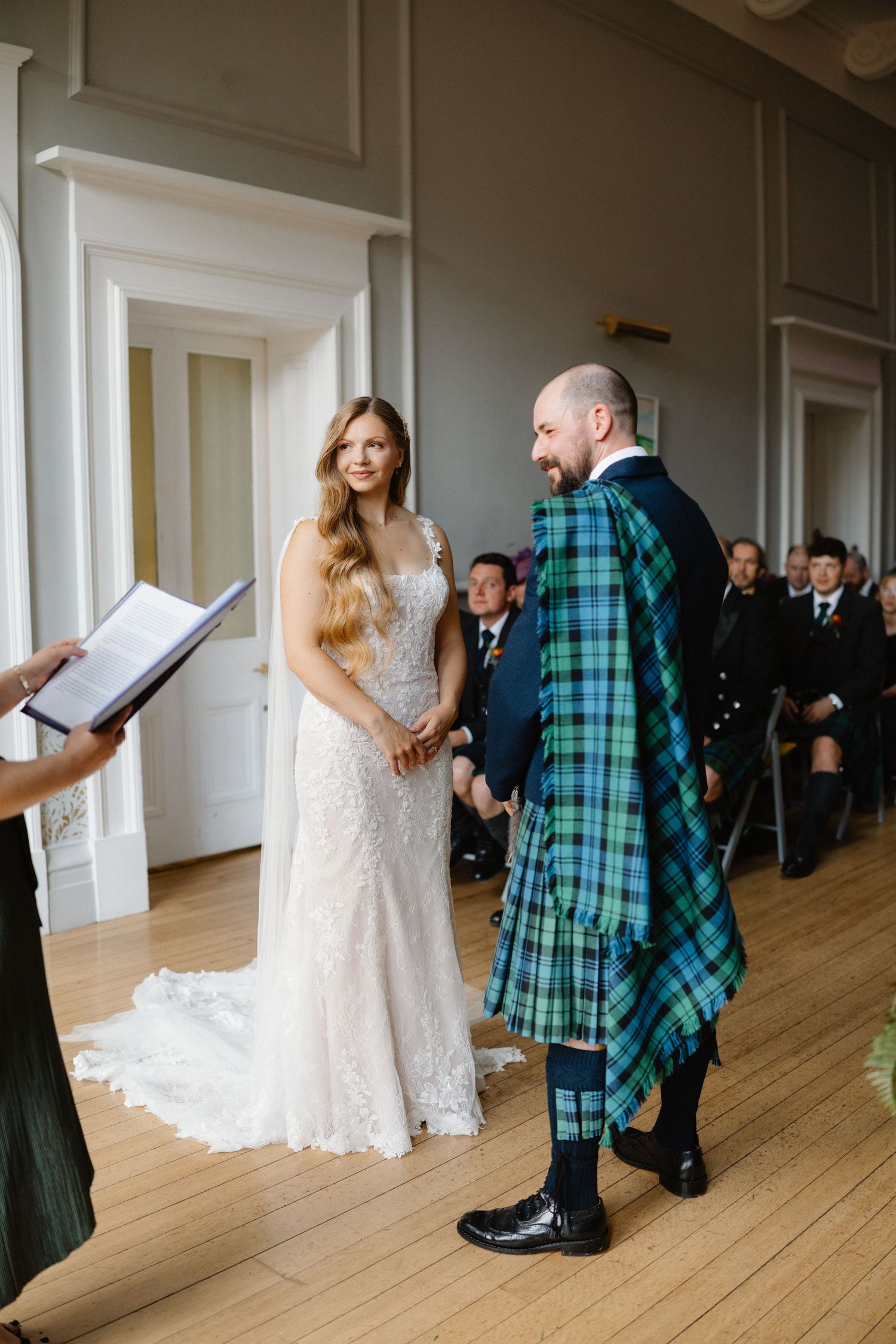 Bride and groom holding hands during wedding ceremony, groom wearing blue and green tartan kilt.
