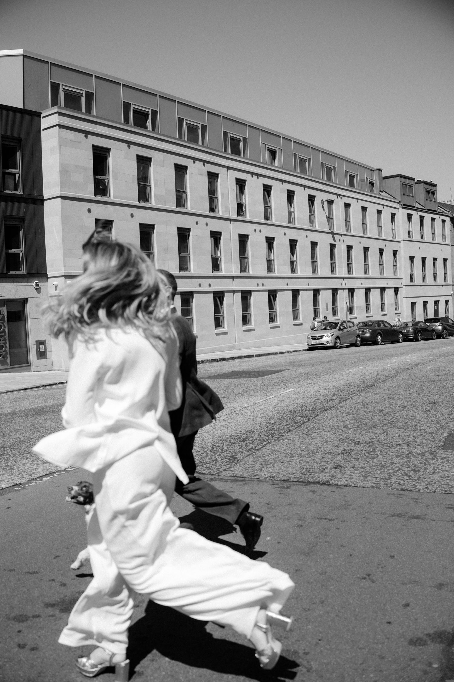 Black and white photo of a bride and groom running down a cobblestone street past a large stone building.