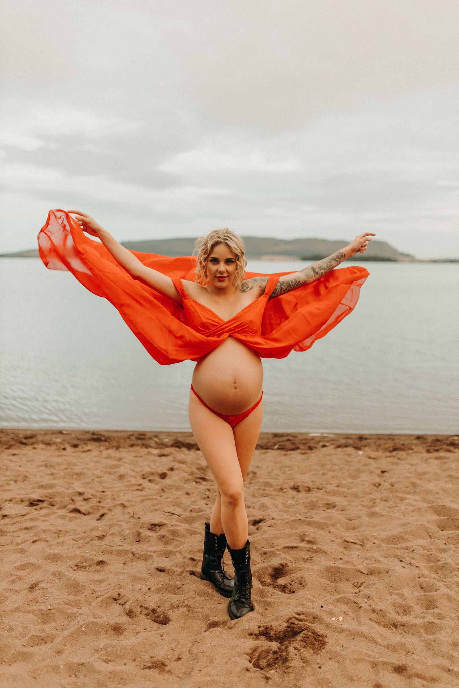 Figure in flowing orange fabric dancing on beach against cloudy sky.