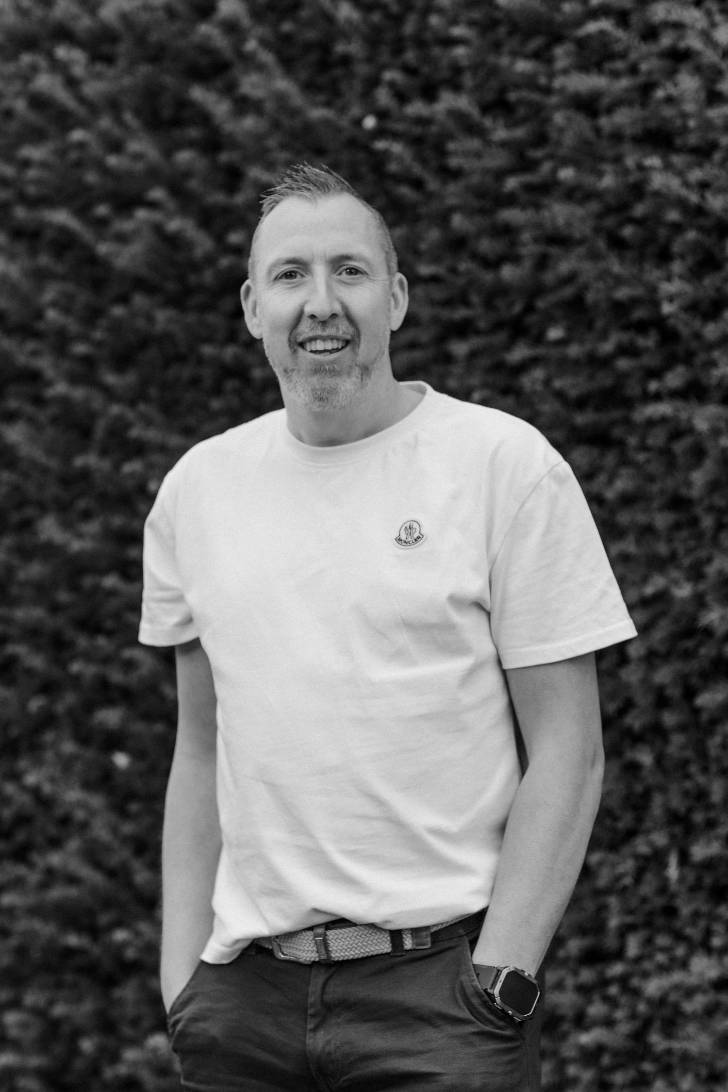 Portrait of a person in a white t-shirt smiling against a hedge background.