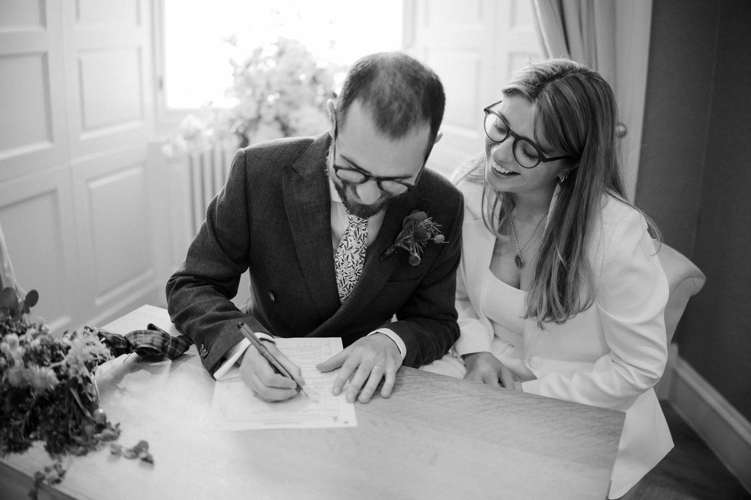 A couple signs marriage documents together at a white desk with flowers nearby.