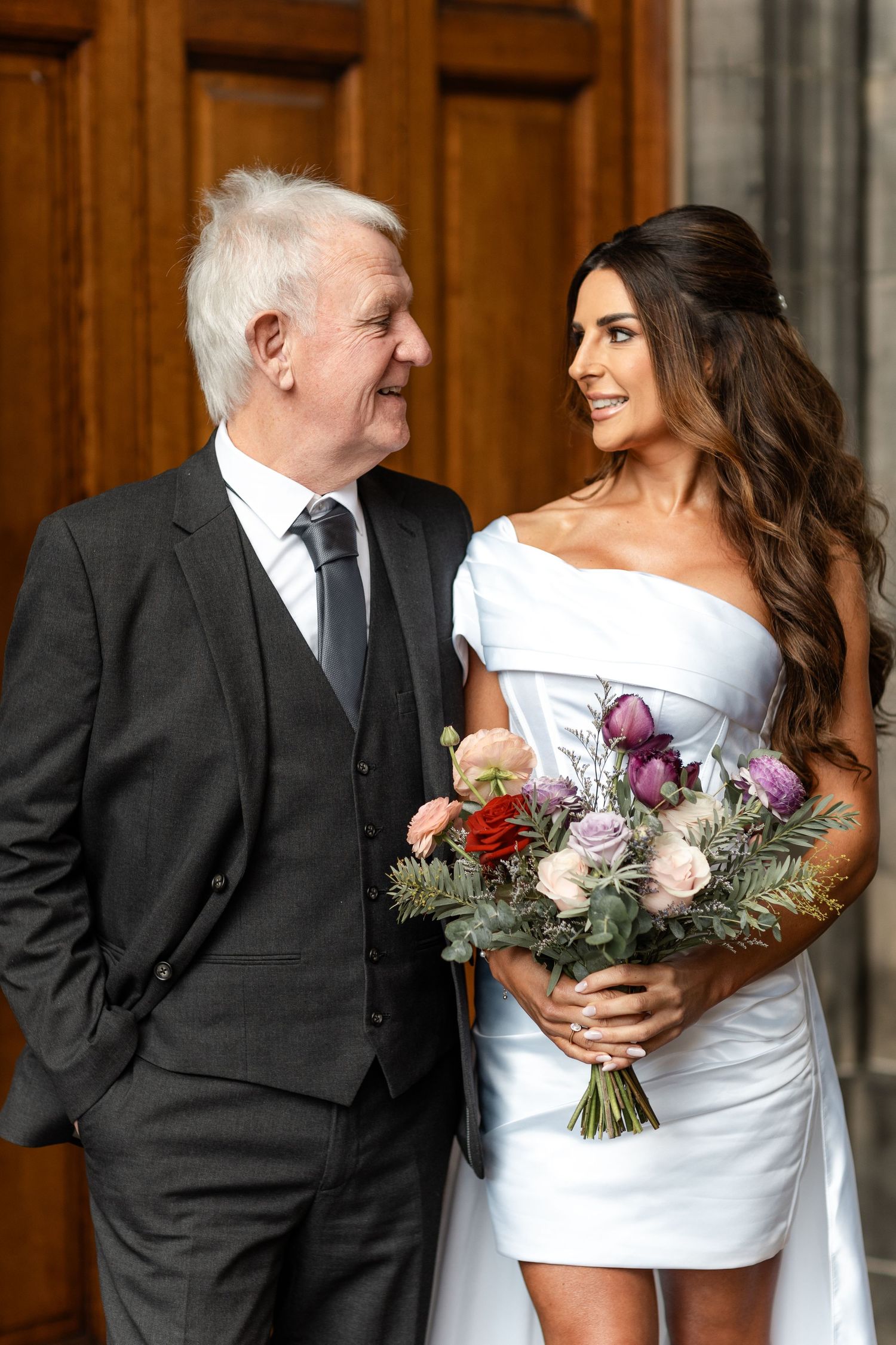 Elegant wedding couple in formal attire posing with floral bouquet against wooden paneled wall.