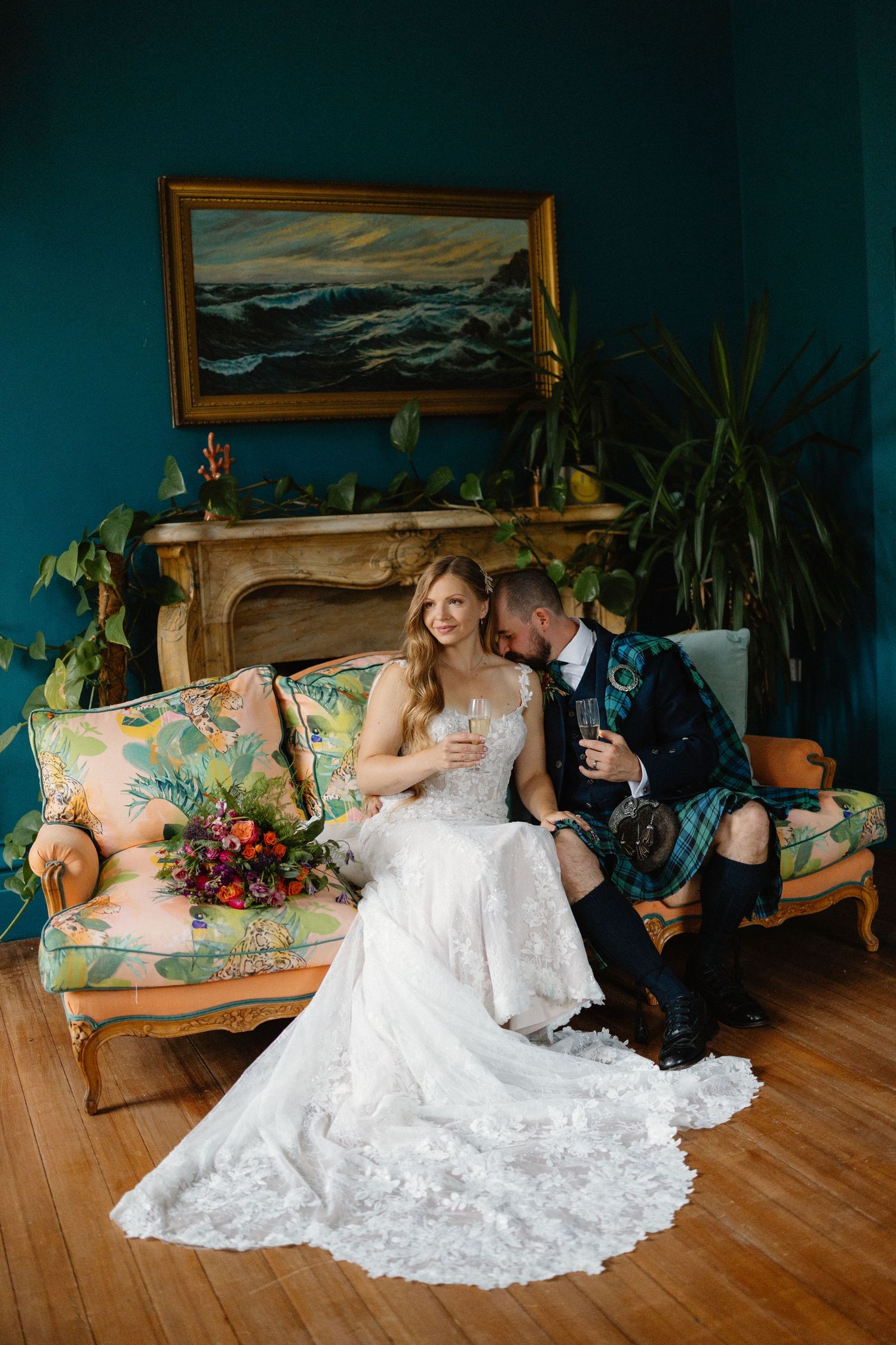 A romantic wedding couple poses on a floral couch against a dark teal wall with ocean artwork.