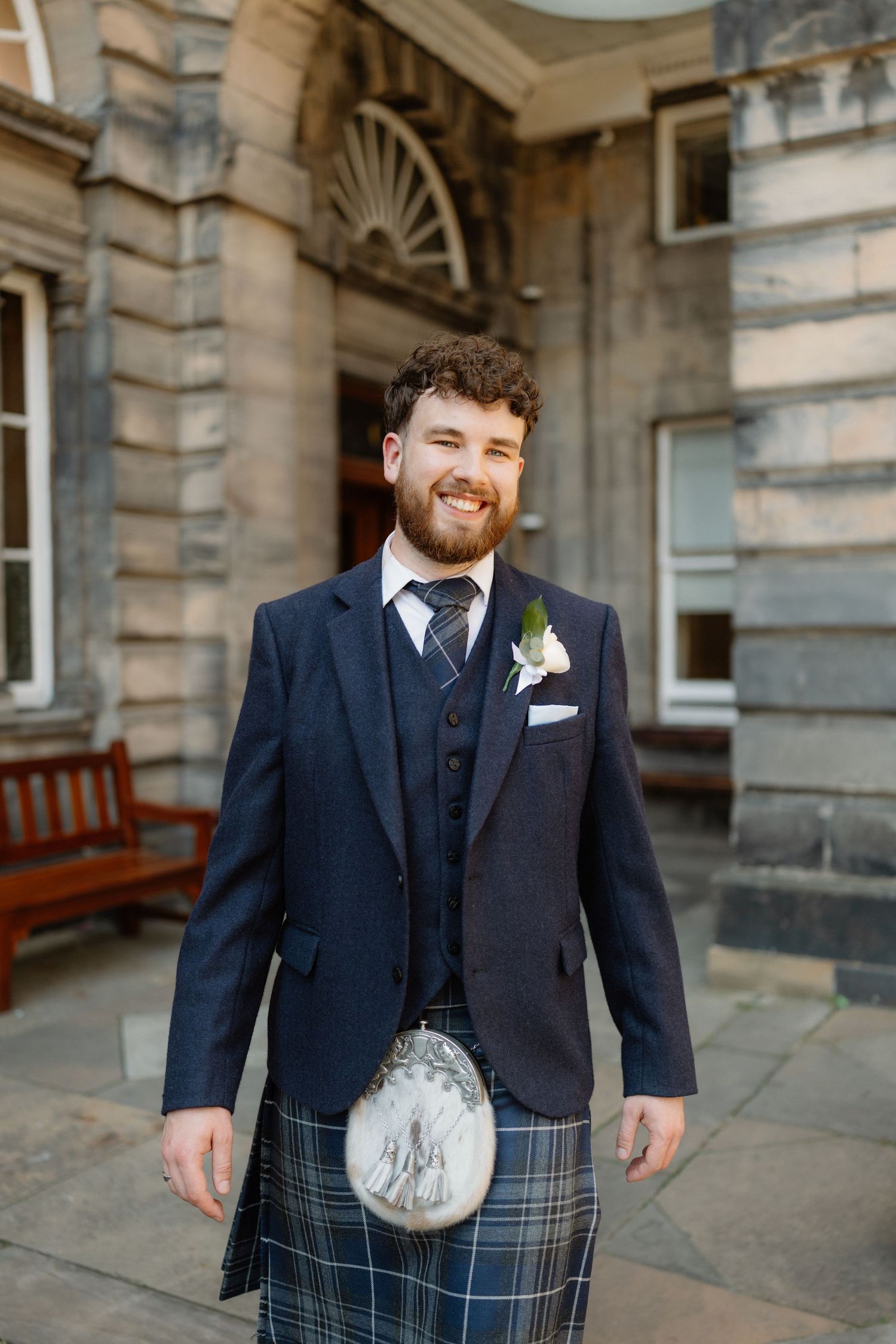 Smiling groom in navy suit, tartan kilt, and floral boutonniere poses outside a historic stone building.
