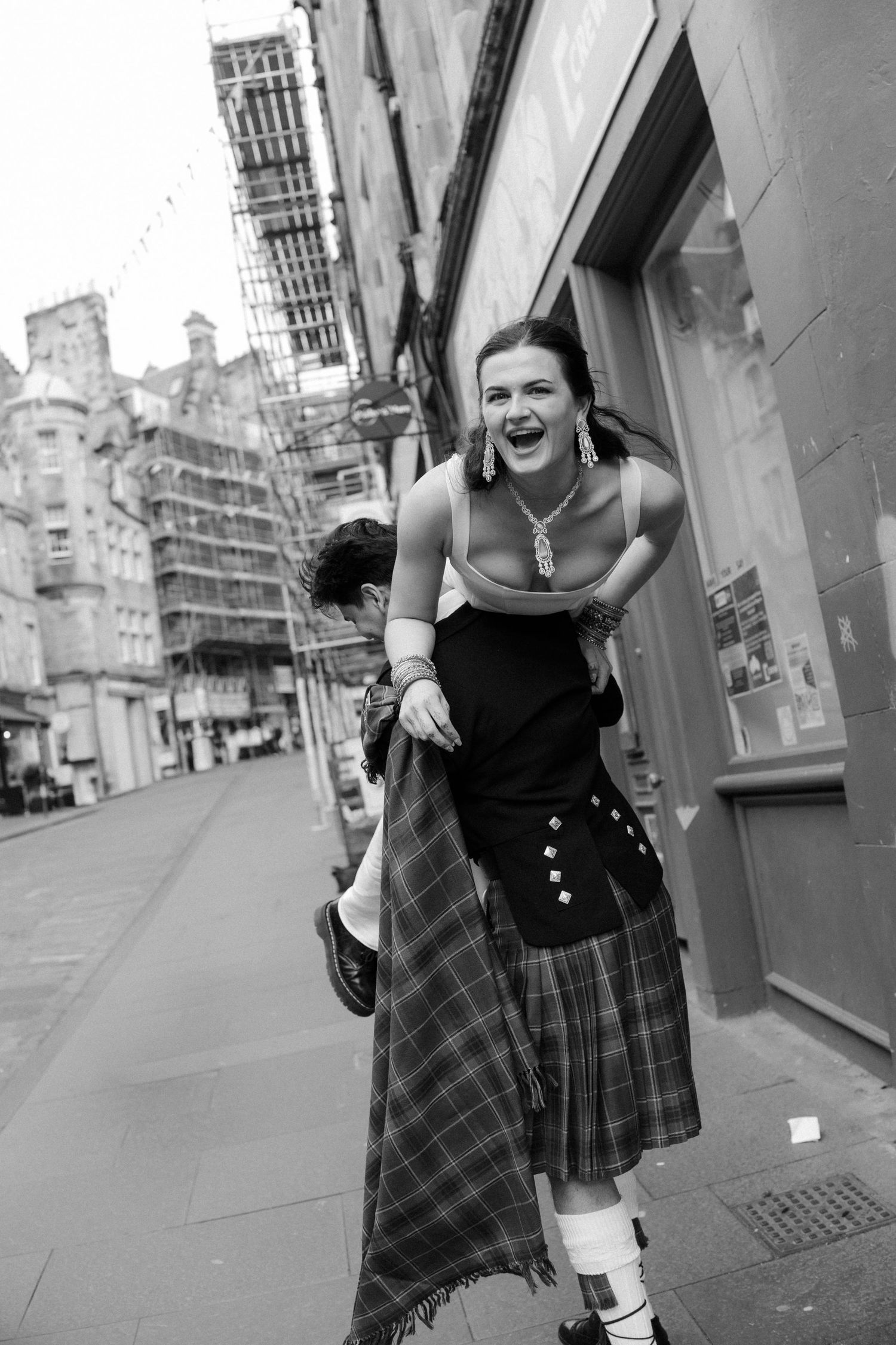 A laughing woman in a plaid skirt and cast on her leg being carried on a mans back on a city street.