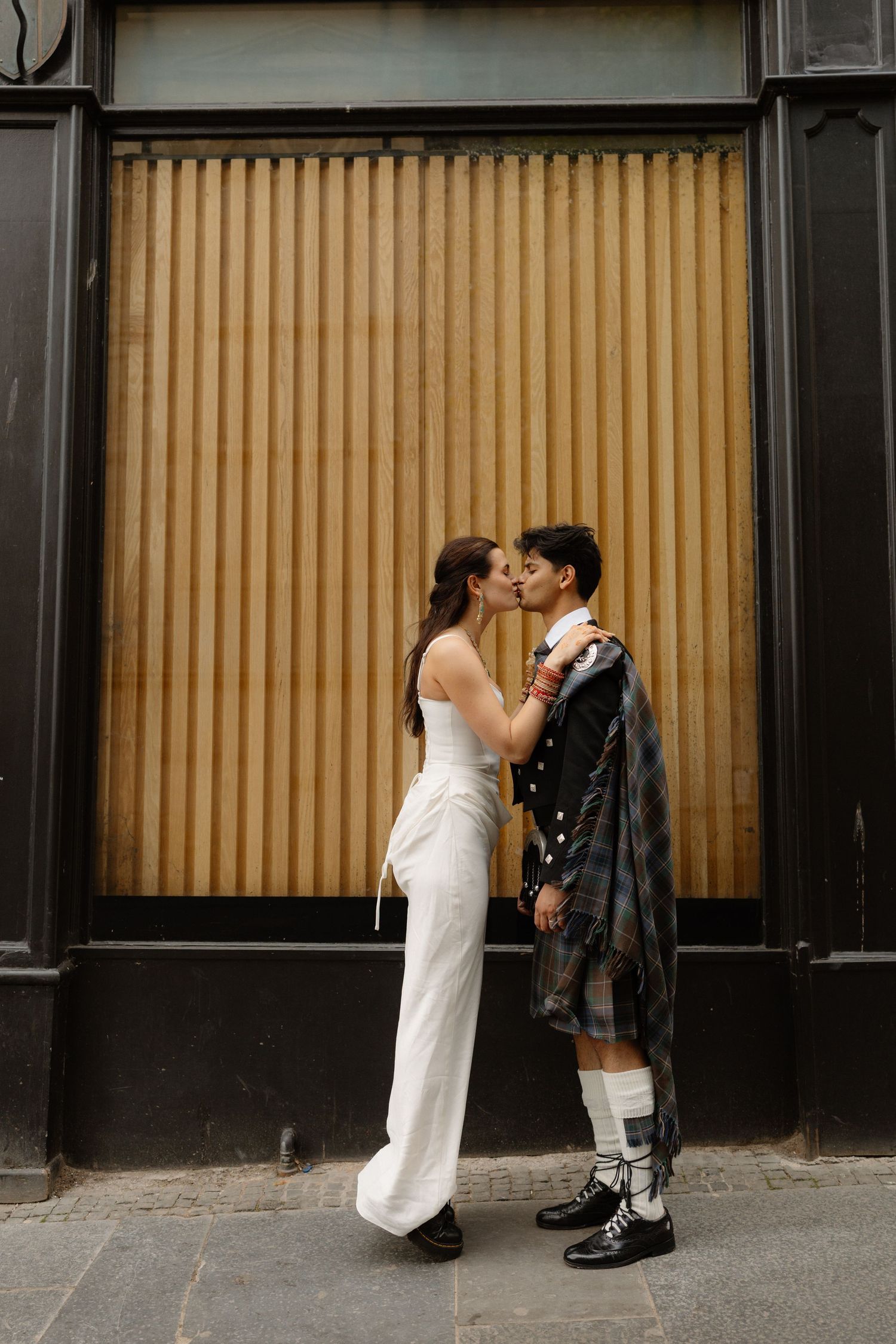 Newlywed couple sharing a romantic kiss on a cobblestone street, groom wearing a Scottish kilt.