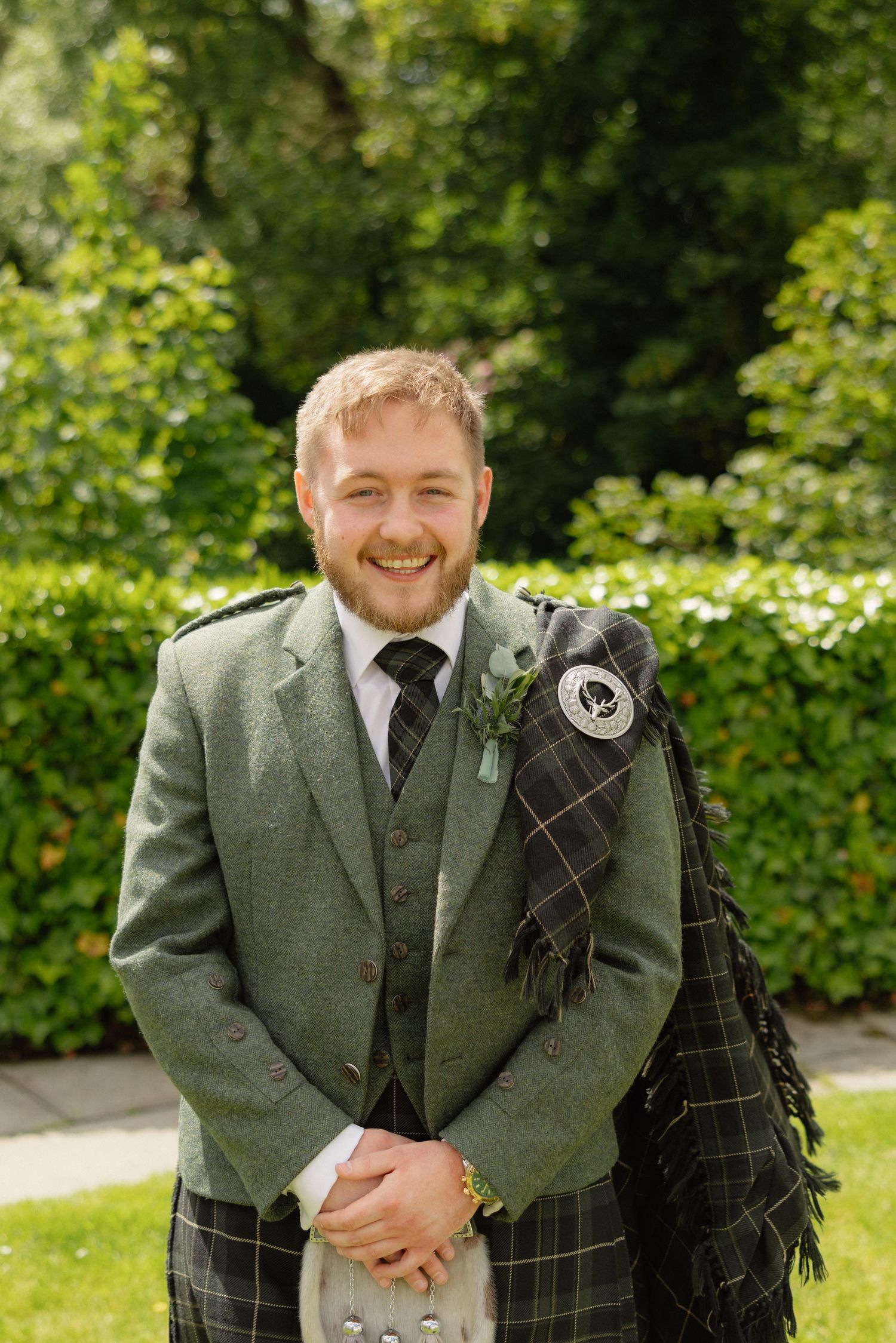 Smiling groom in green tartan kilt and jacket with boutonniere standing outdoors at Scottish wedding.