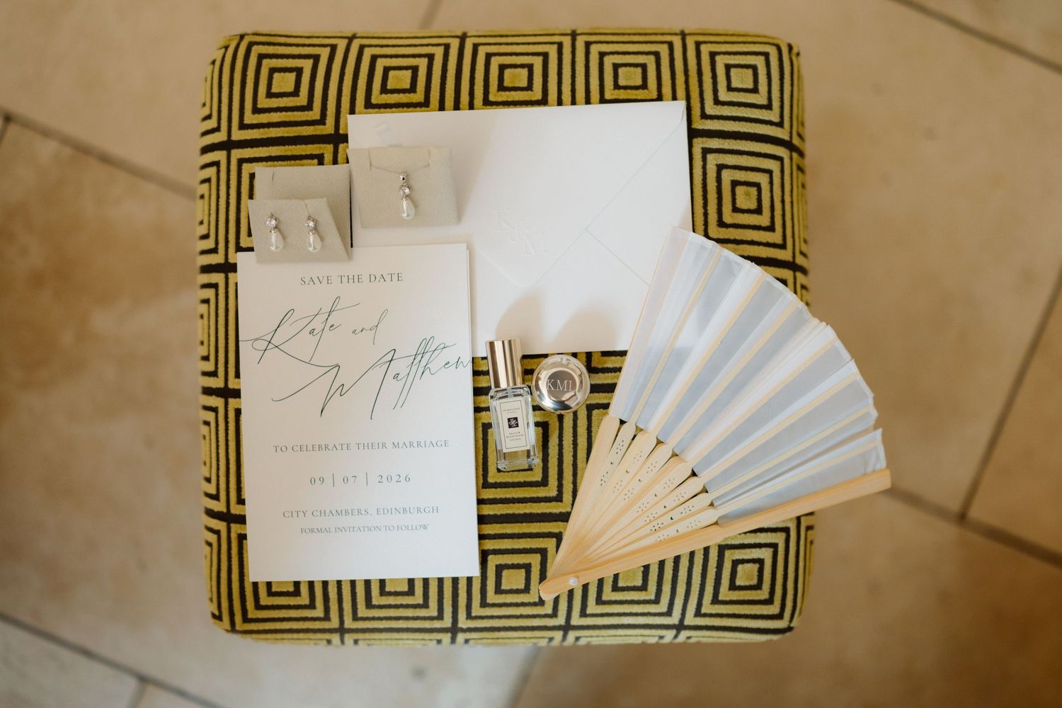 Wedding stationery flat lay with save the date card, white paper fan, and small photos on a gold geometric patterned ottoman.