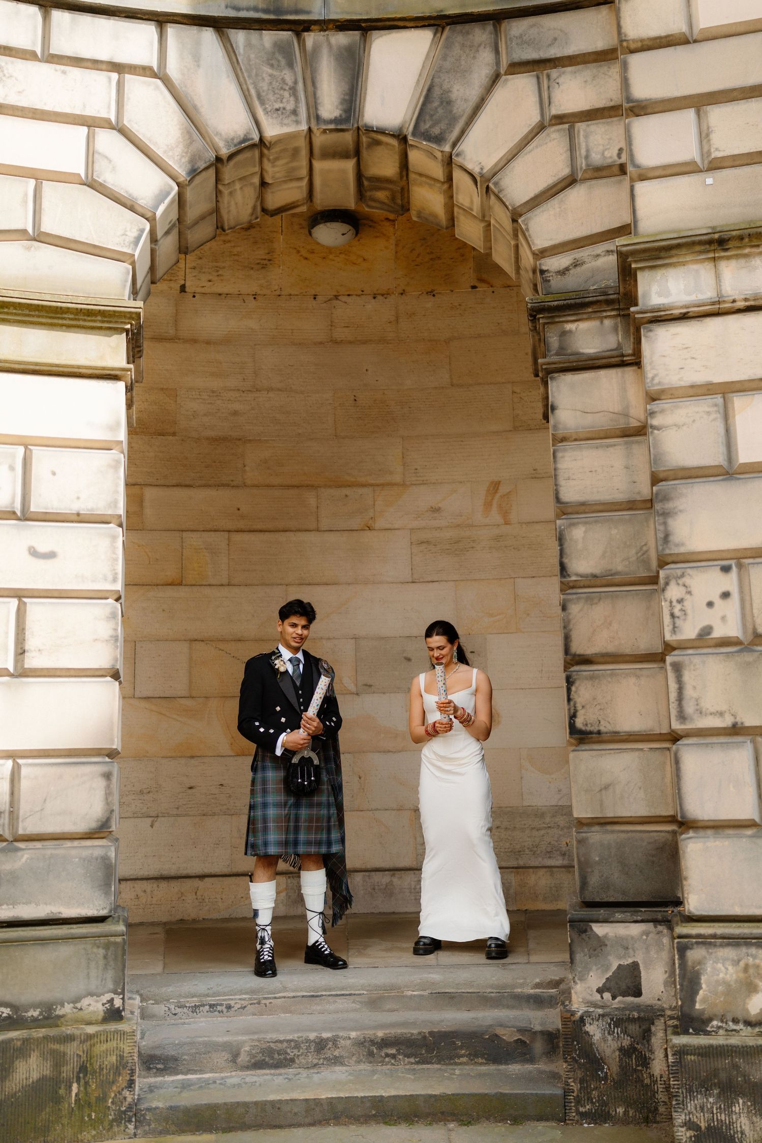 Bride in white gown and groom in Scottish kilt standing beneath a stone archway at a historic Edinburgh venue.