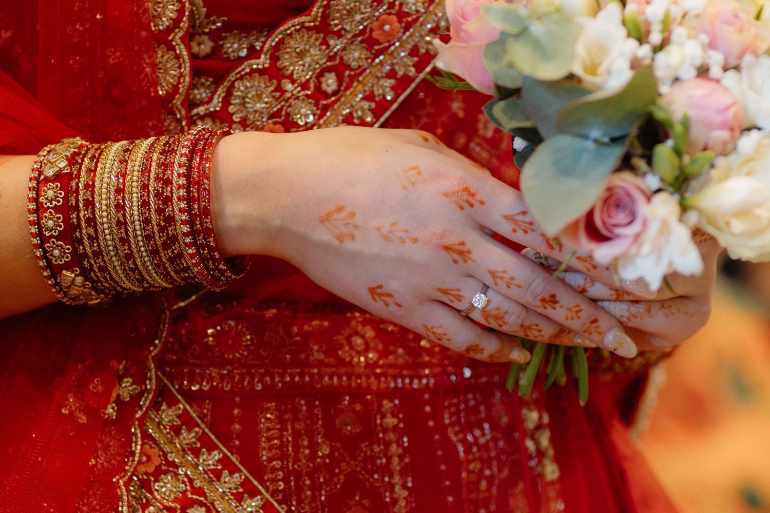 Bride in red saree with henna mehndi designs on hands, gold bangles, holding pink and white wedding bouquet.
