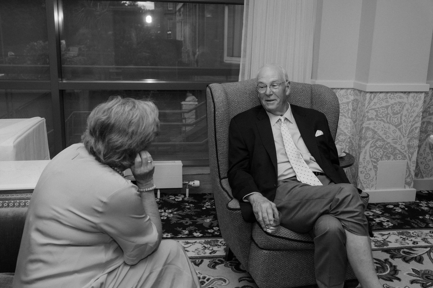 Black and white photo of an elderly man in a suit conversing with a woman in a cozy armchair setting.