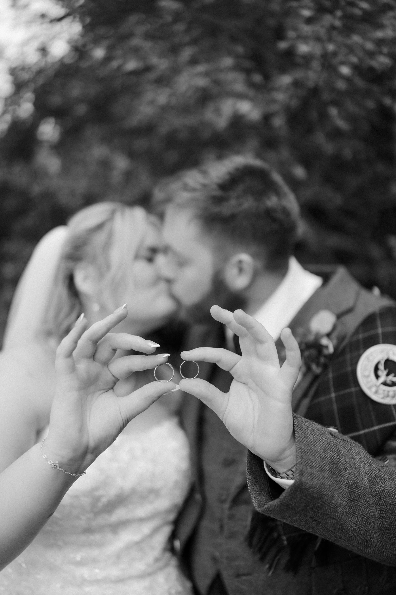 Black and white photo of newlyweds kissing while holding up their wedding rings outdoors.