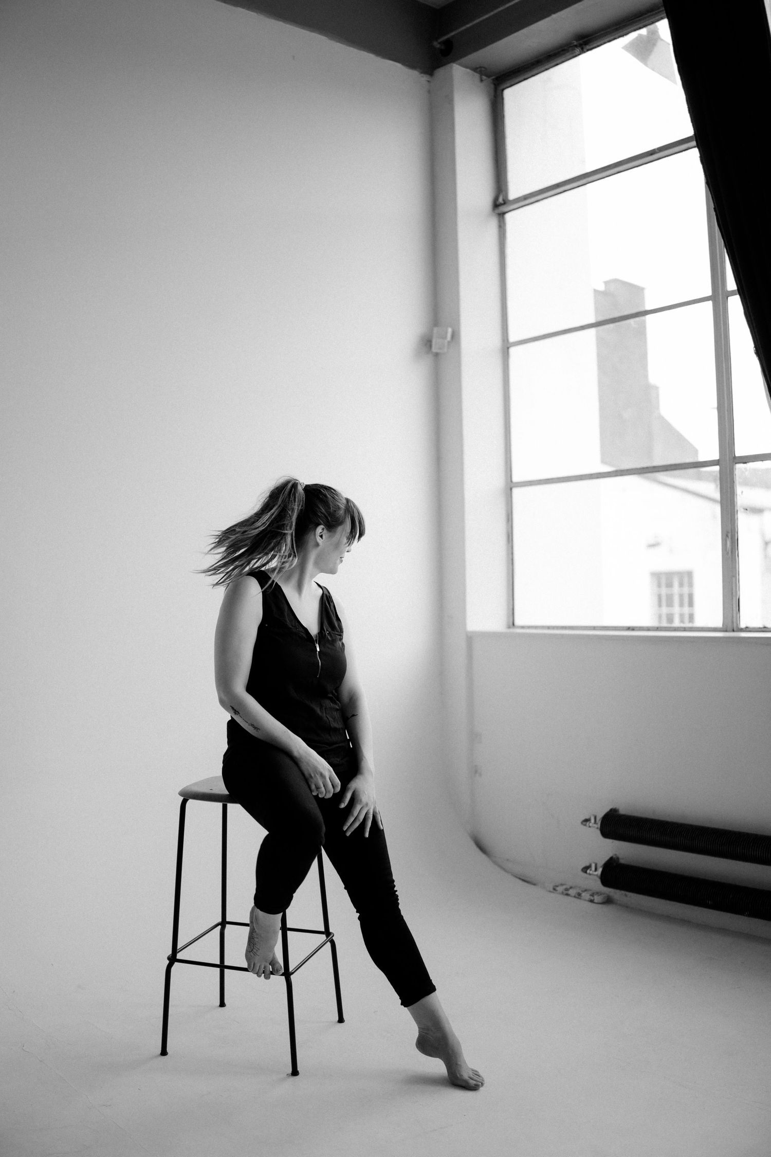 Monochrome photo of dancer posing on stool near window in studio setting.