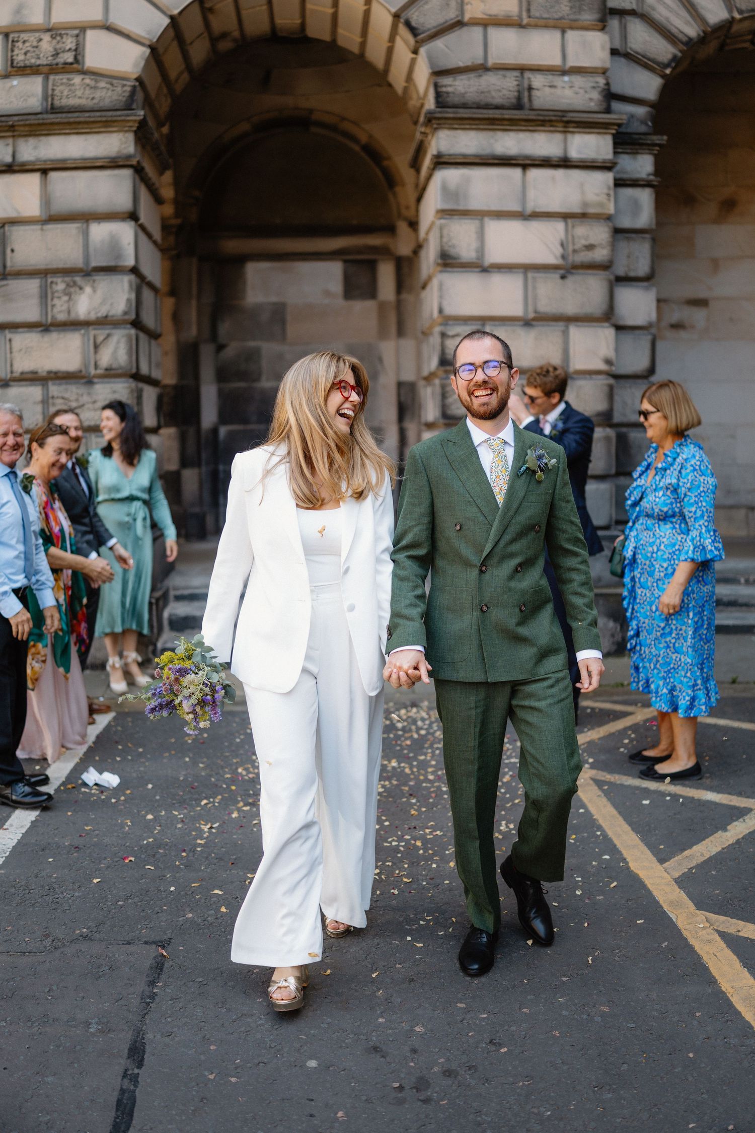 Wedding party walks through stone archways while guests throw confetti in celebration.