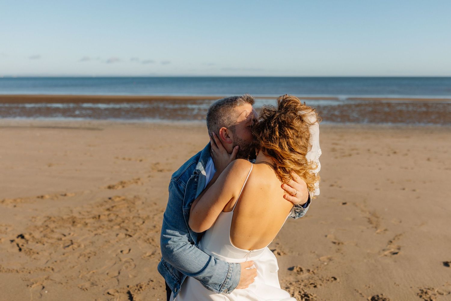 Romantic couple embraces on a sandy beach under blue skies.