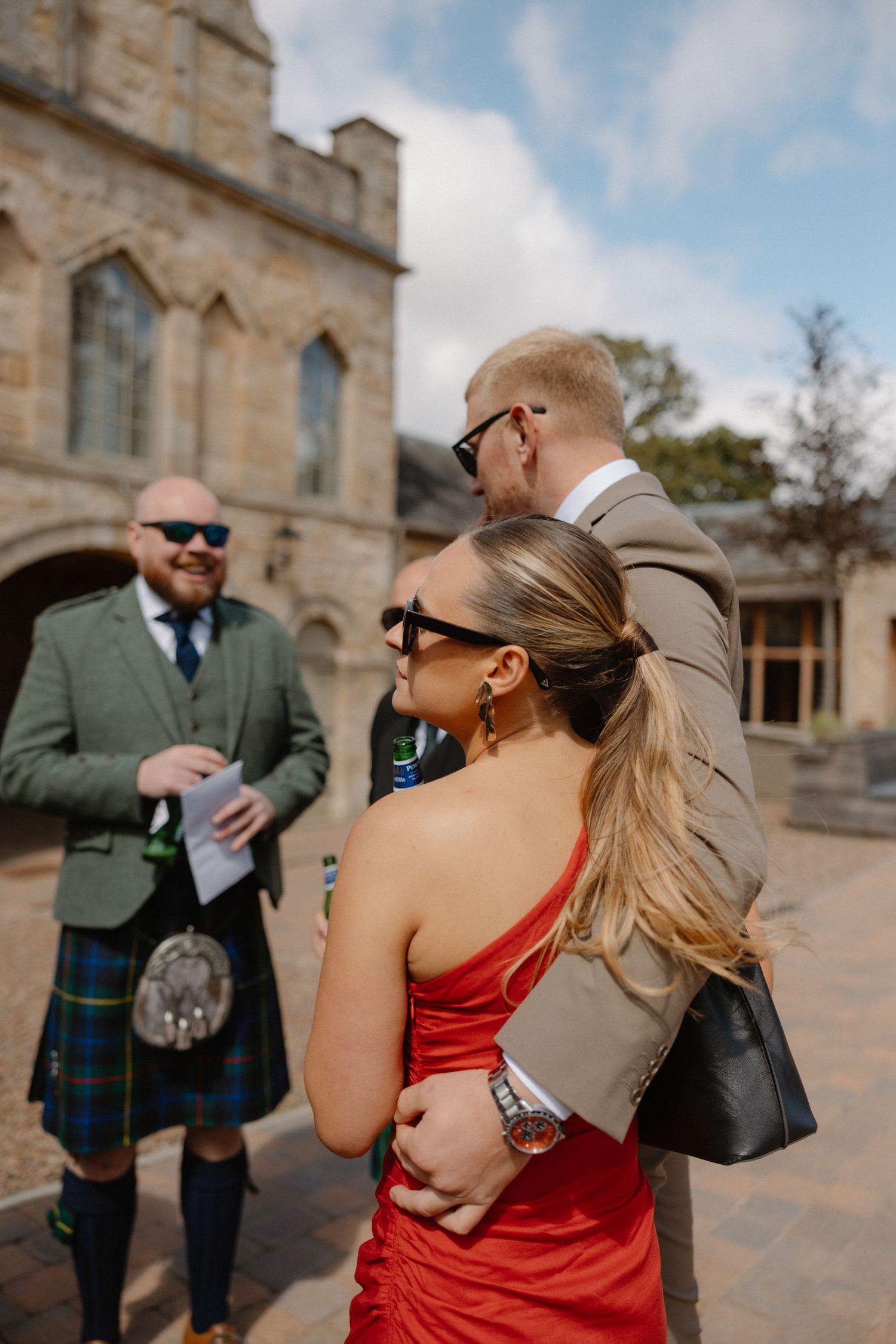 Wedding guests in kilts and formal attire gather outside stone building.