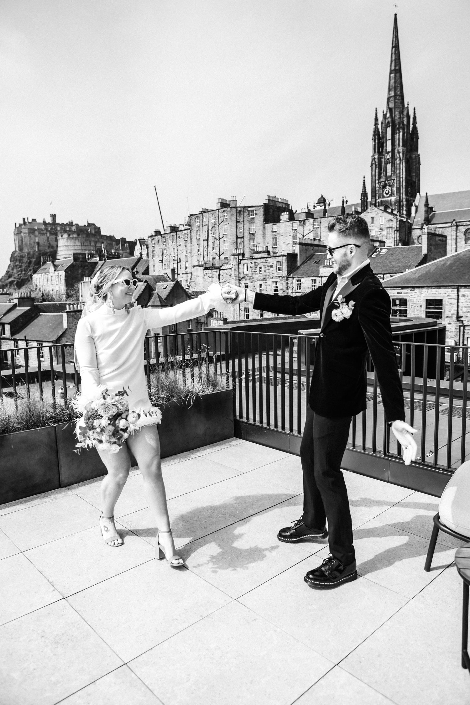 A couple dances playfully on a rooftop terrace with Edinburgh's historic skyline and church spires in the background.