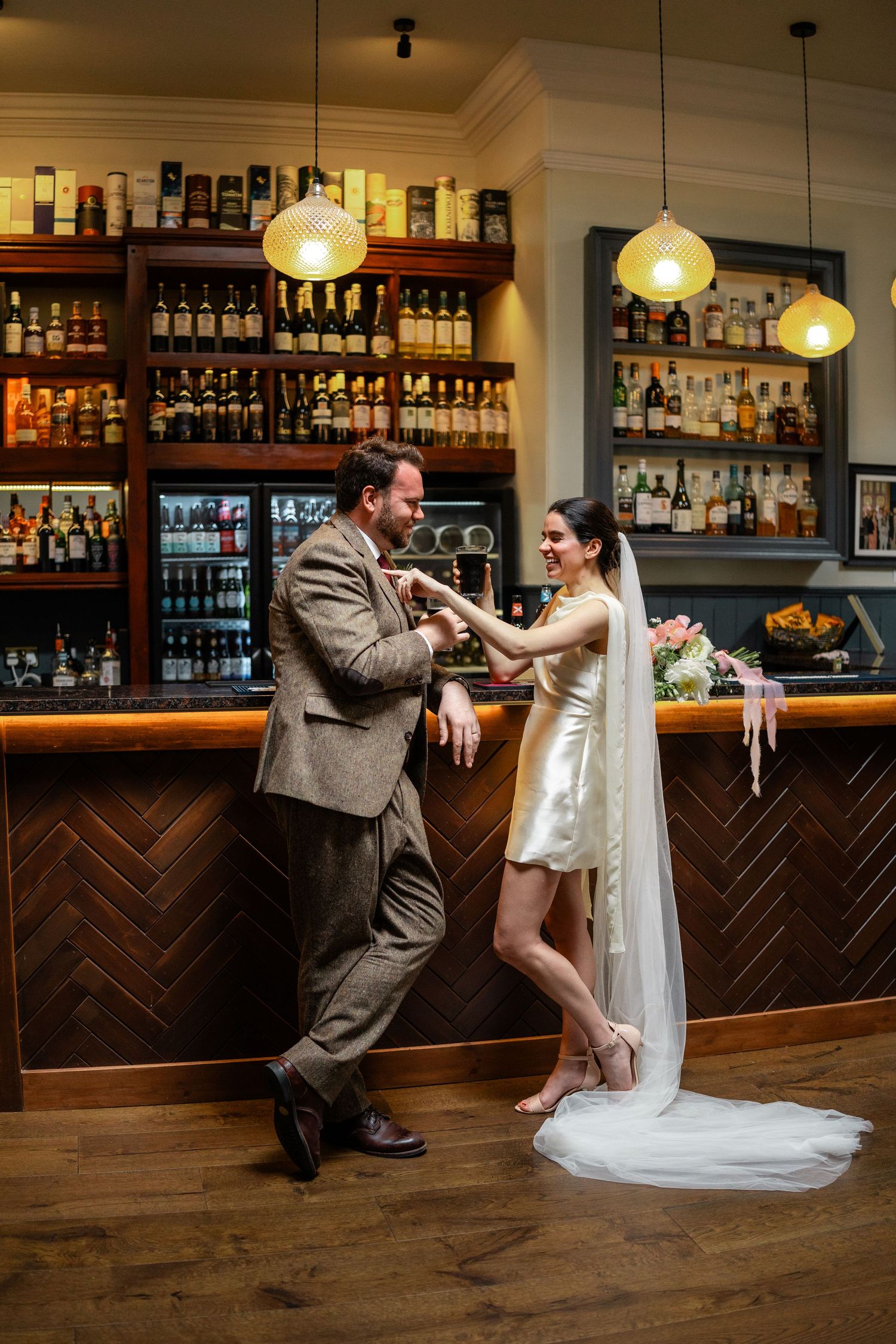 Playful wedding couple dances and poses in front of a wooden bar with bottles displayed.
