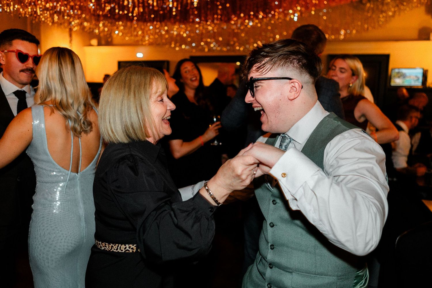 Wedding reception guests dancing and celebrating under warm lighting and decorative string lights.