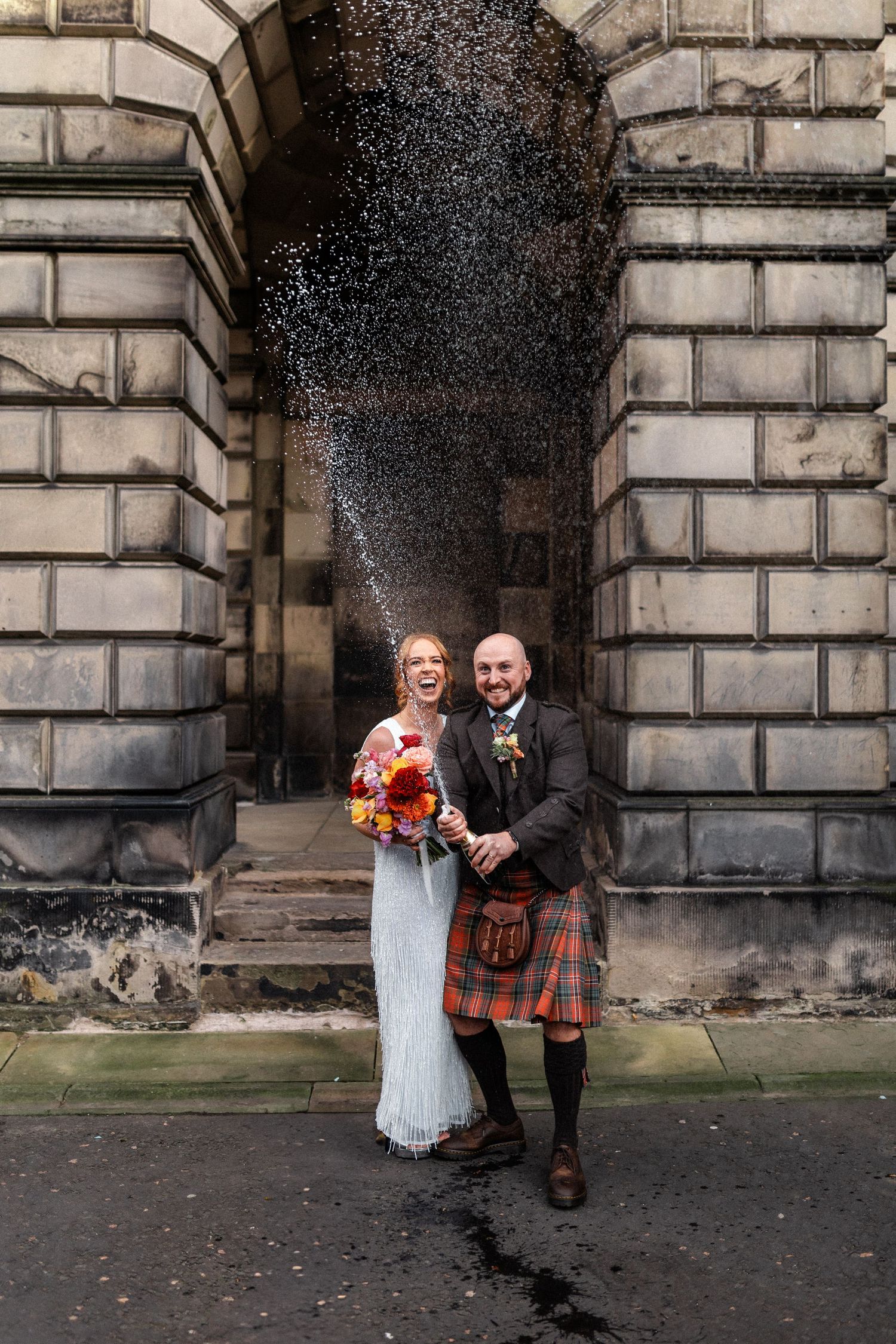 Couple shares intimate moments against stone architecture backdrop in Scottish wedding attire.