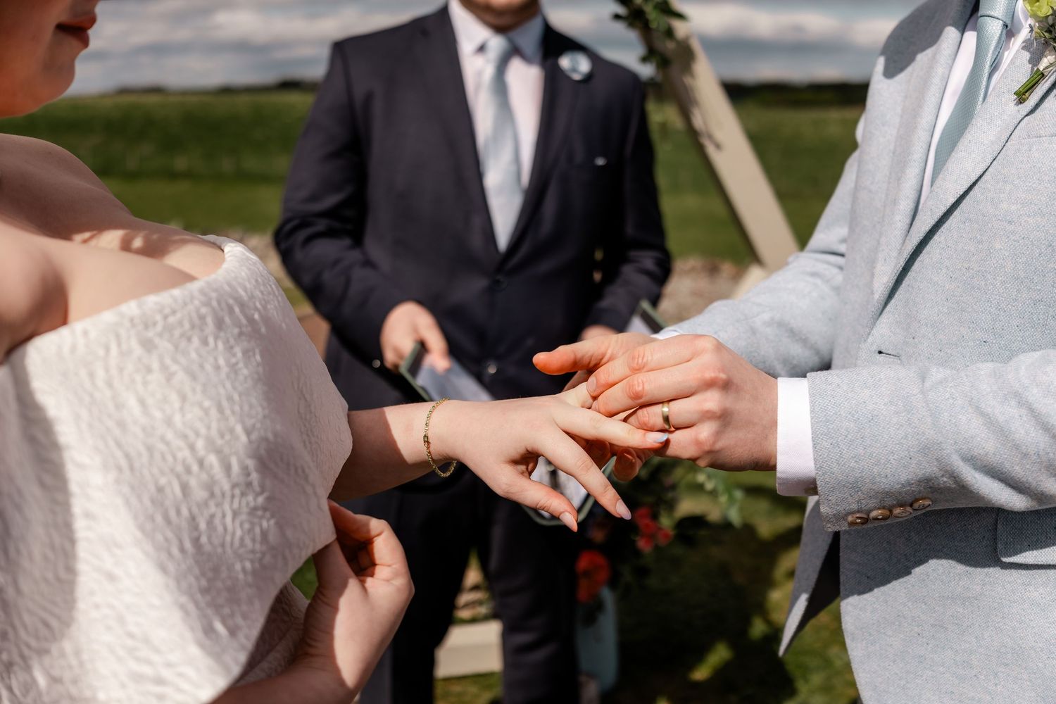 Close-up shot of wedding rings being exchanged during an outdoor ceremony against a cloudy sky.