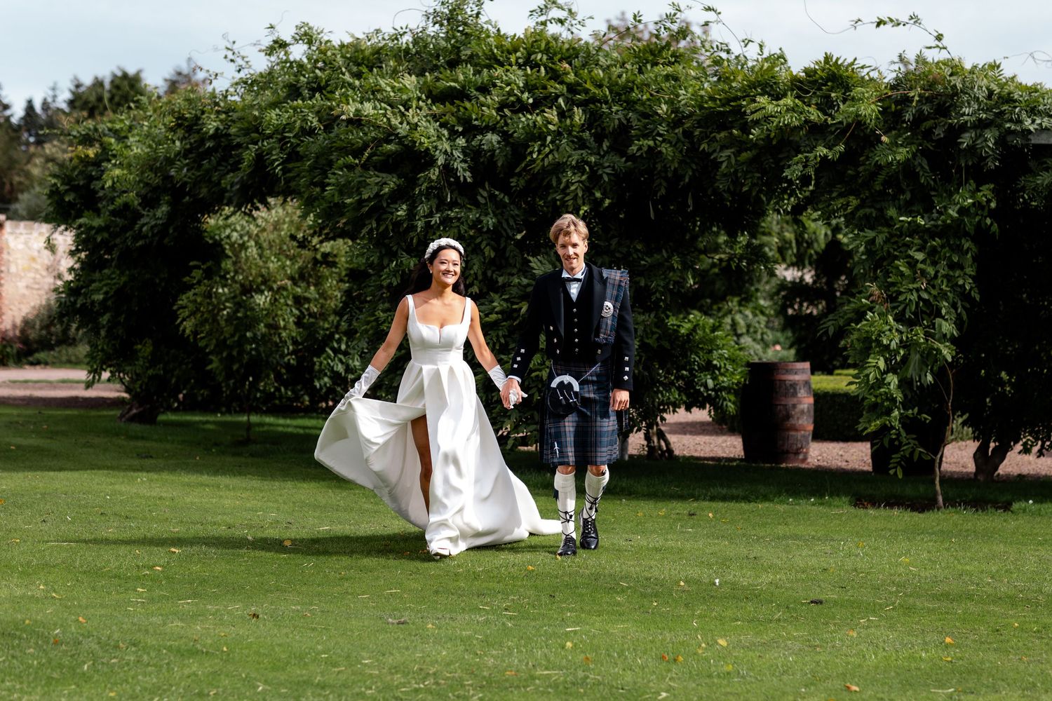 A couple walking across a manicured lawn with traditional Scottish wedding attire.