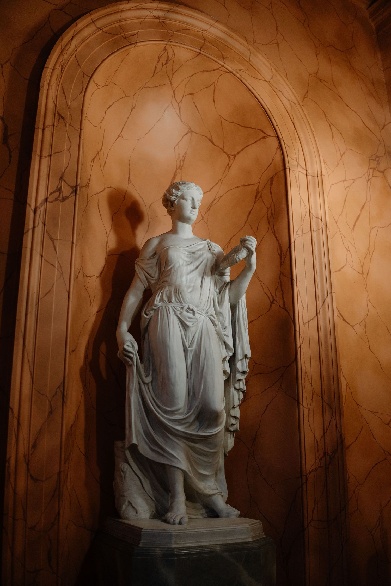 Classical marble statue in a draped robe stands elegantly within an arched alcove with textured marble walls.