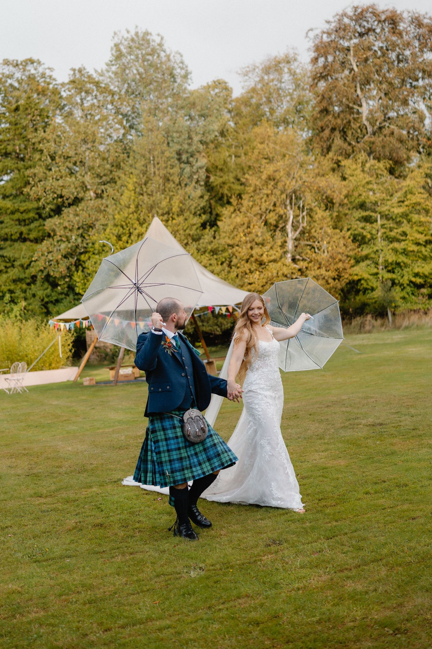 A couple dances playfully with clear umbrellas in a grassy field surrounded by autumn trees.