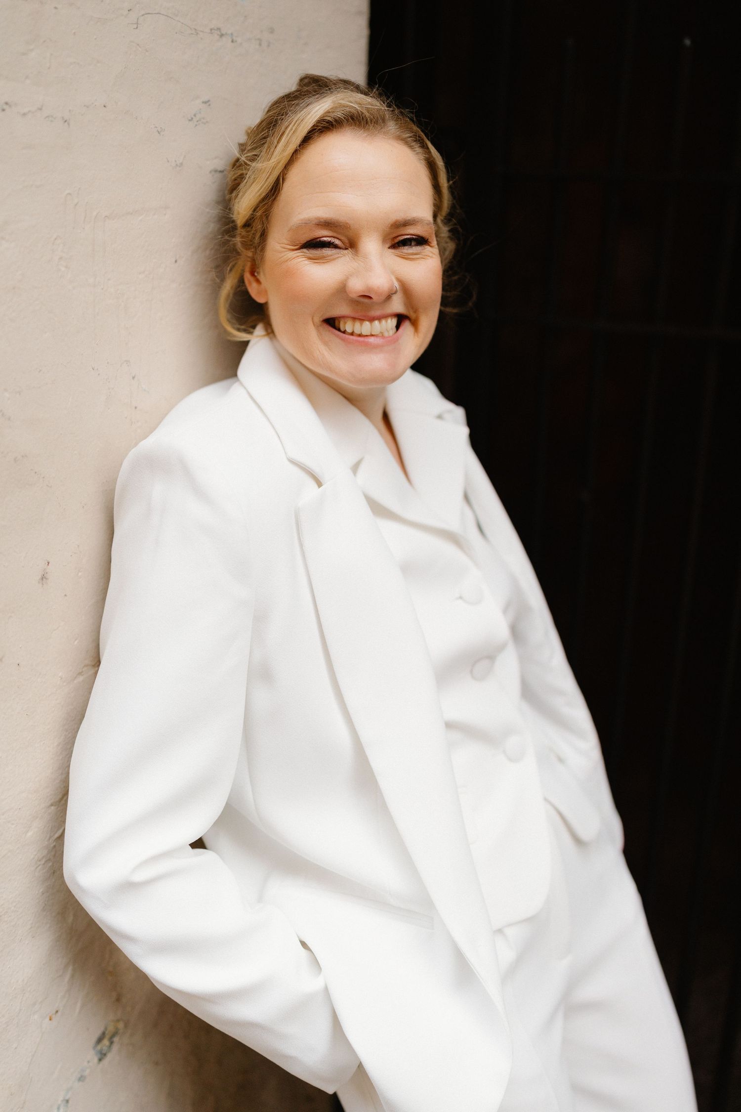 Smiling woman in a white blazer suit leaning against a wall, exuding elegance and confidence.