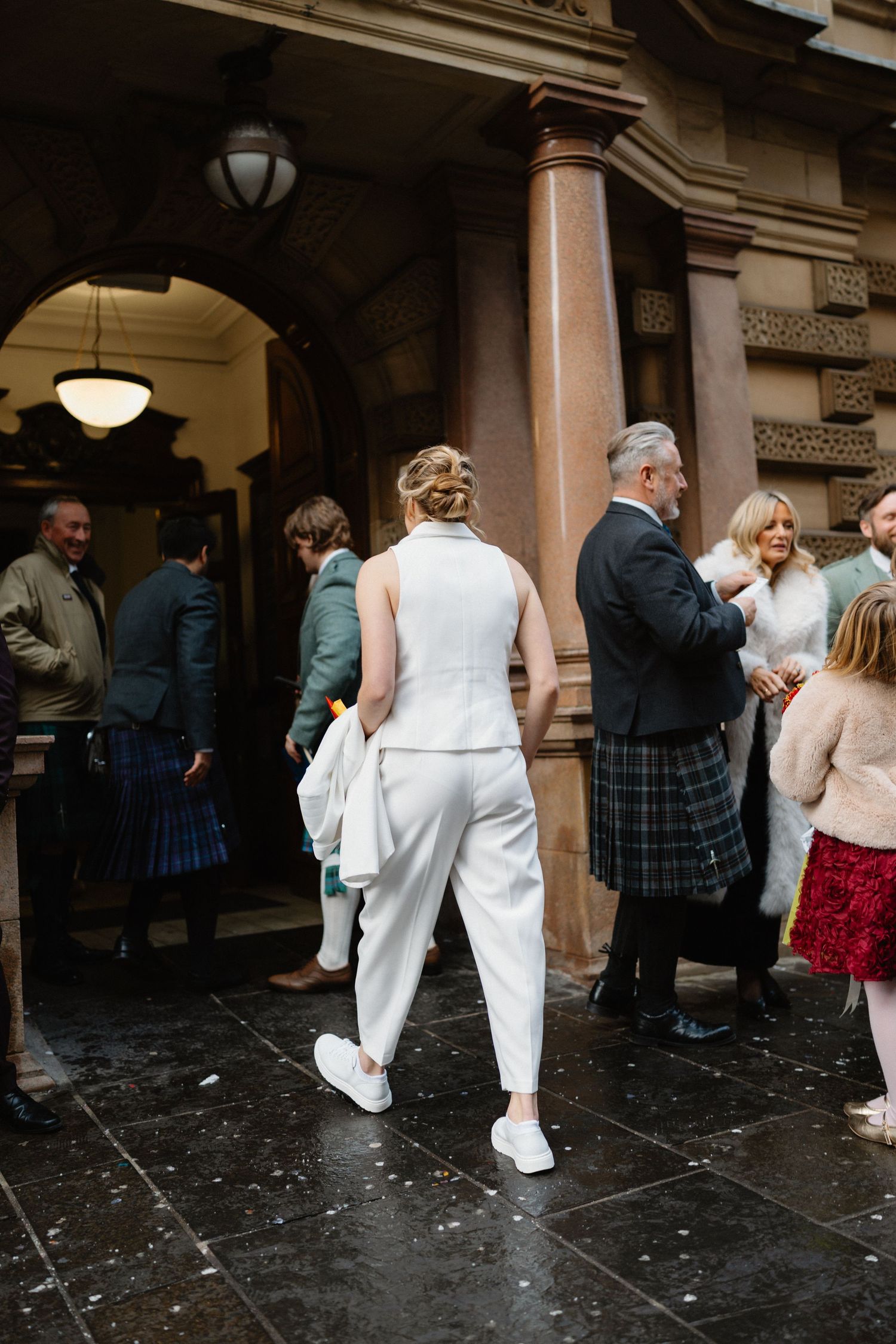 Bride in white trouser suit and sneakers walks toward wedding venue entrance with guests in kilts and formal wear.