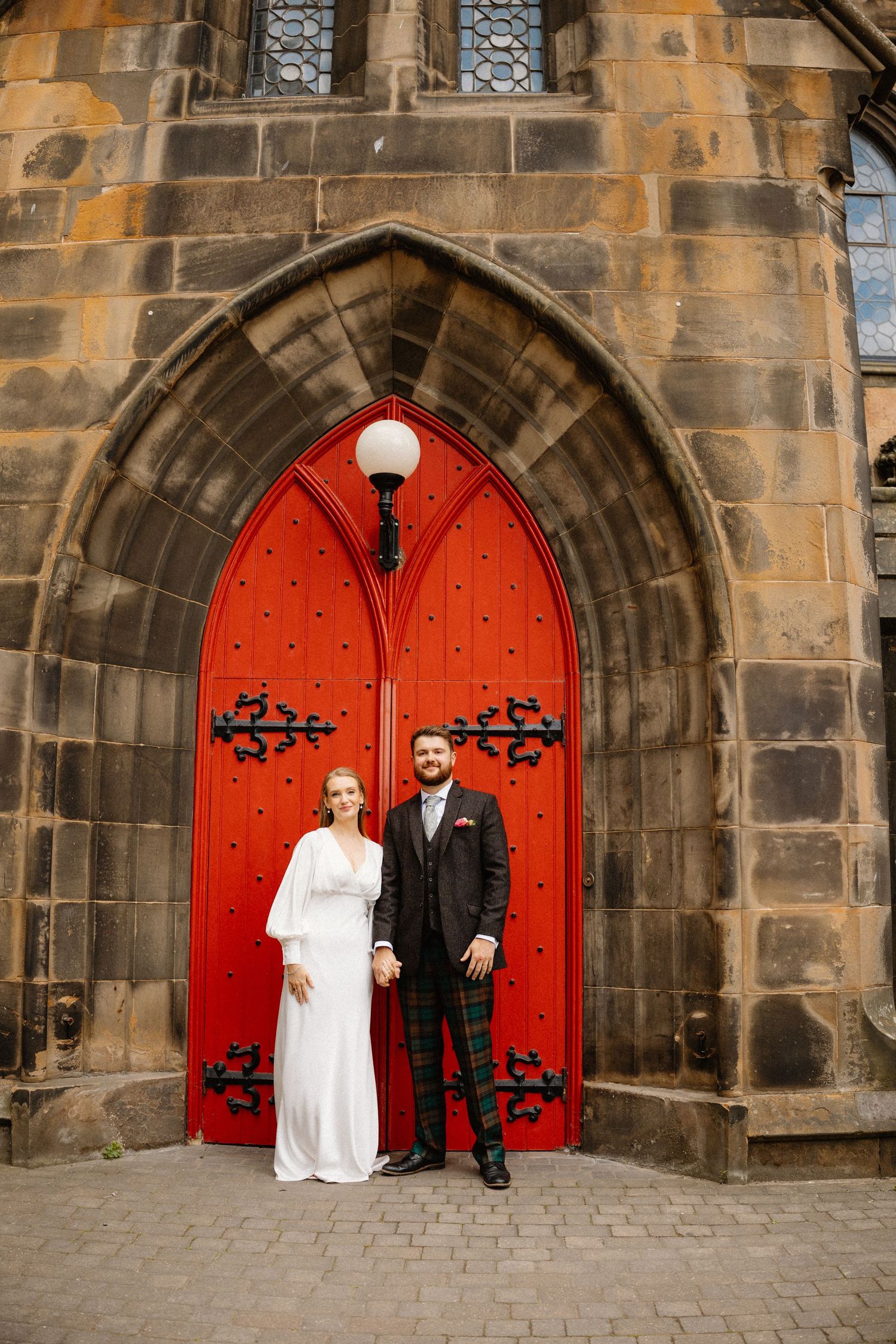 Bride and groom standing before a stunning red Gothic arched door of a stone church on their wedding day.