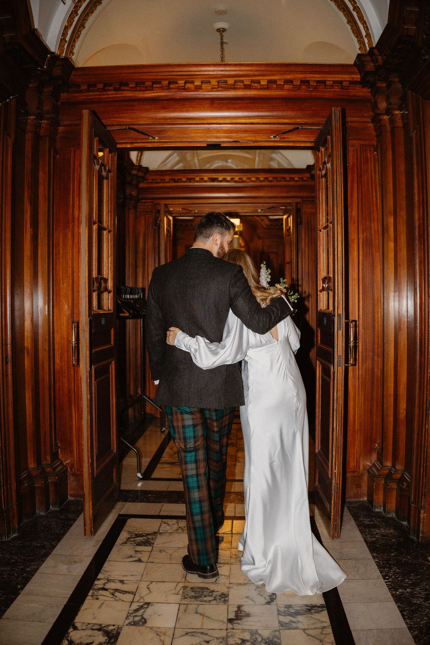 Bride and groom walk arm in arm through ornate wooden doorway in a historic building.