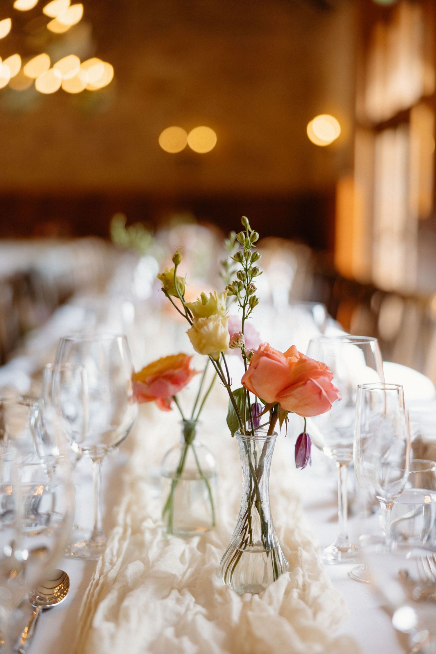Elegant wedding reception table with pink and yellow roses in glass bud vases on a white ruffled tablecloth.