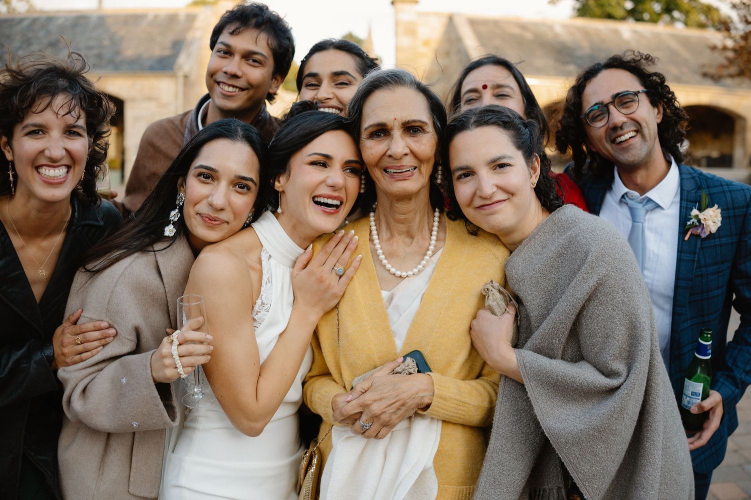 Bride poses with smiling family members and friends in a warm outdoor wedding group photo.
