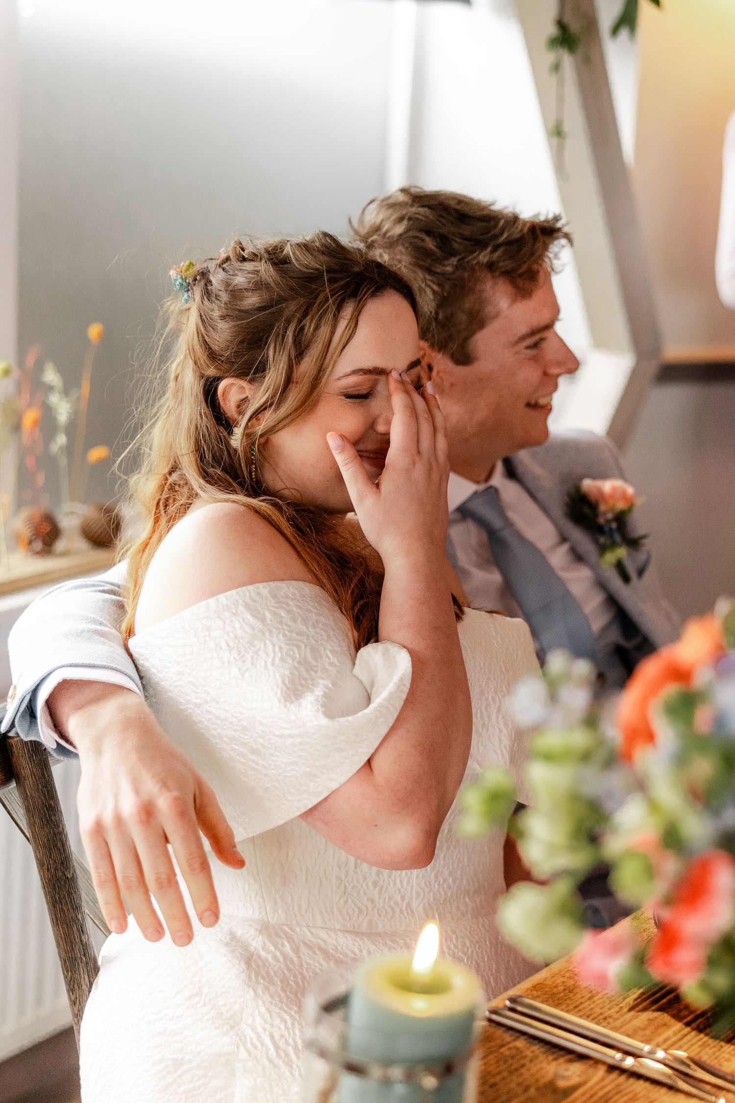 Bride laughing and wiping tears while groom smiles and embraces her at wedding reception table.