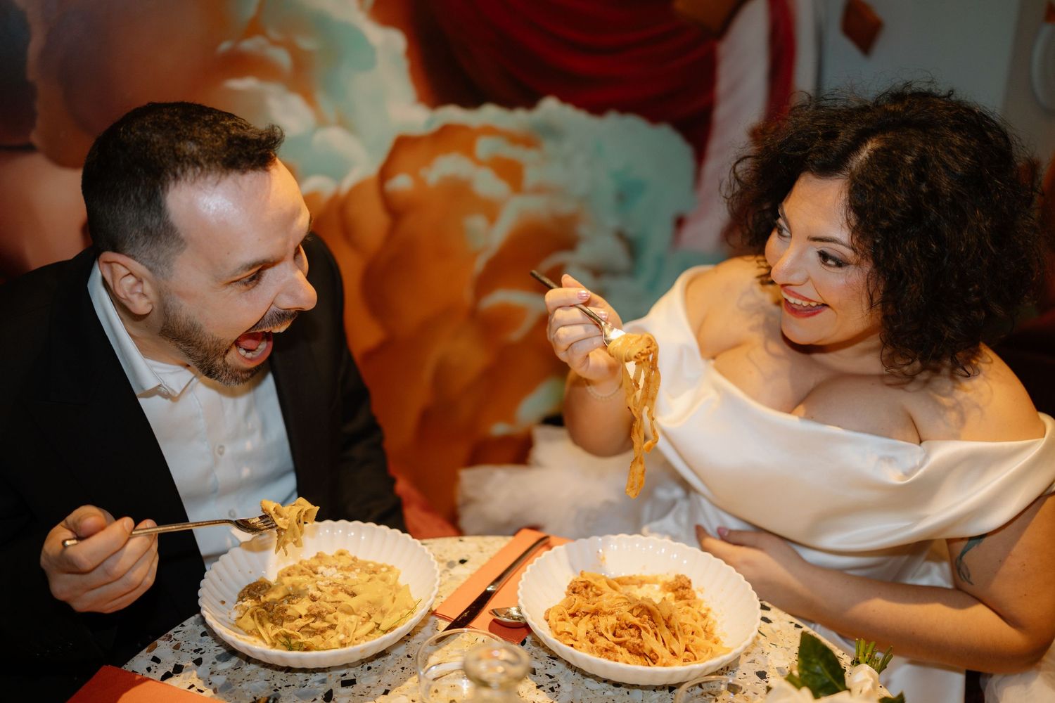 A joyful couple in formal attire sharing pasta dishes at a romantic restaurant, laughing together on their wedding day.