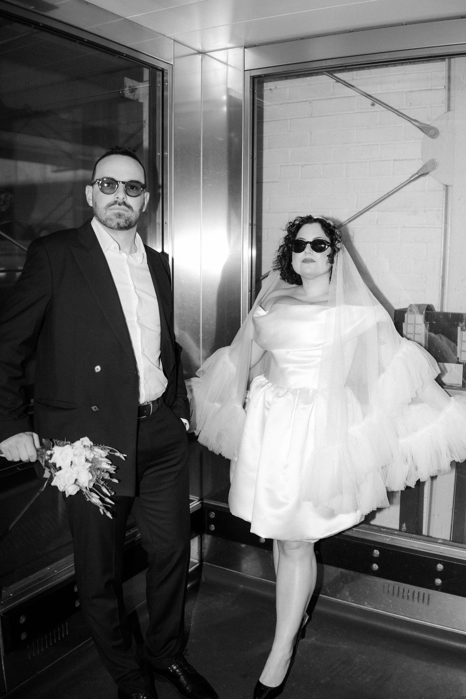 Stylish bride and groom wearing sunglasses posing in elevator doorway in black and white wedding photo.