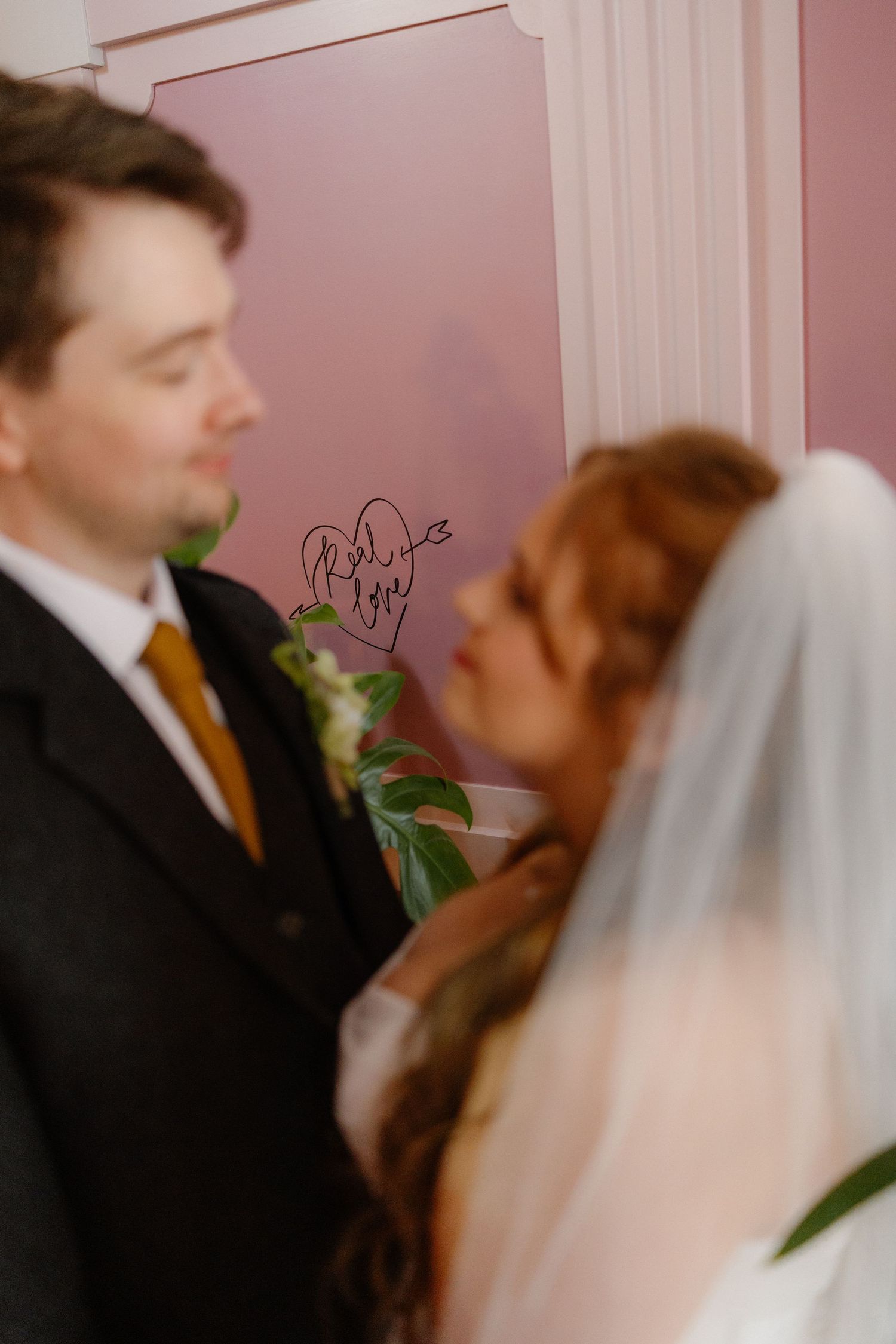 Bride in veil and groom in suit standing before pink wall with heart inscription at wedding venue.
