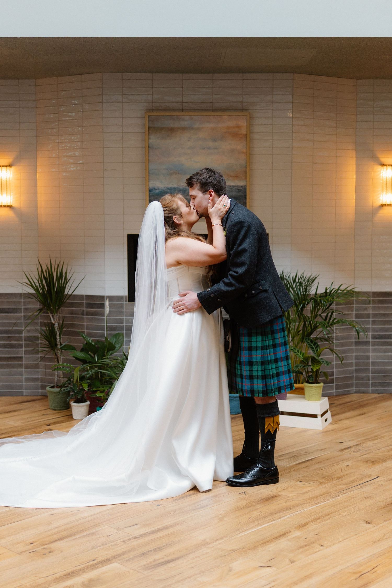 Bride and groom sharing first kiss, groom wearing traditional Scottish kilt on wedding day indoors.