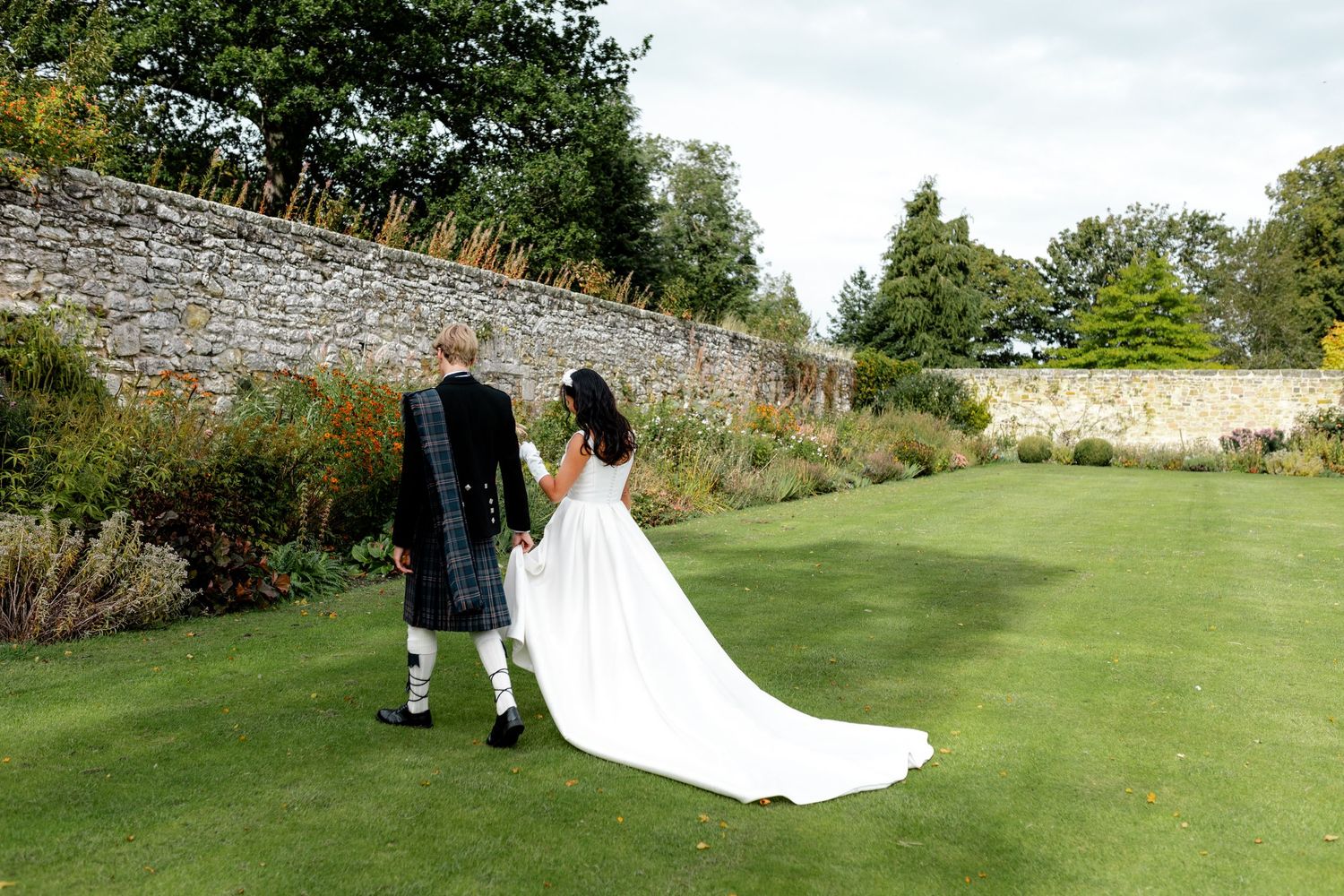 Bride and groom walk hand in hand across lush lawn, wedding dress trailing, beside a stone garden wall.