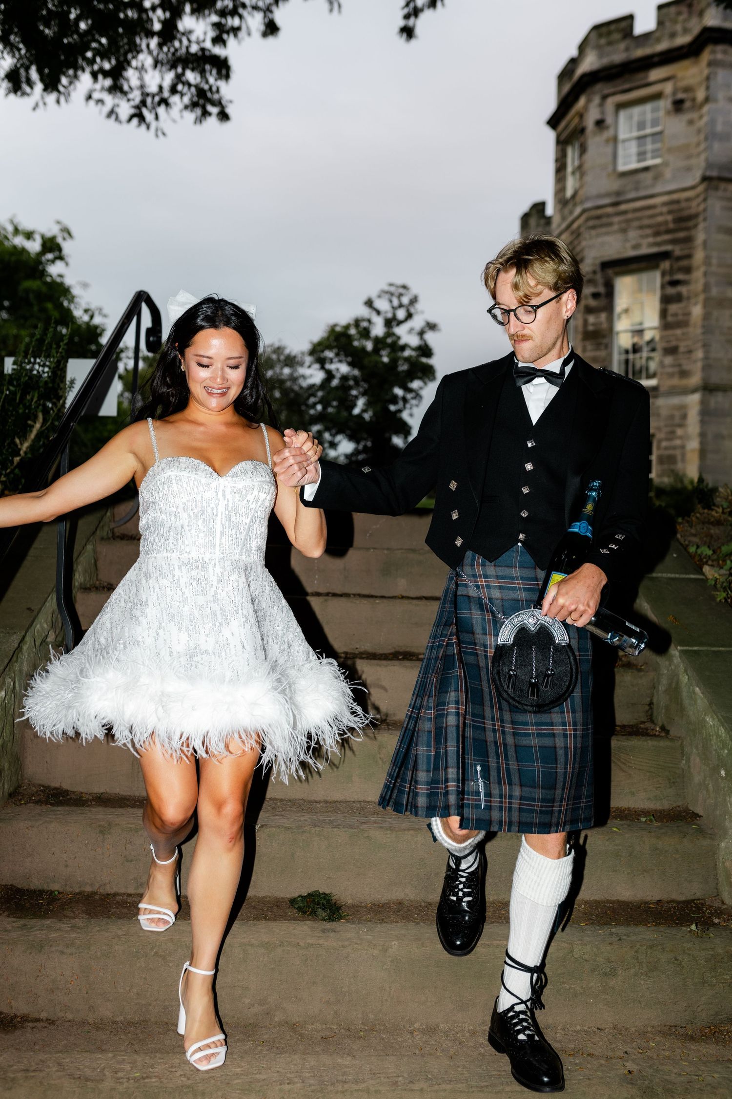 Bride in feathered mini dress and groom in tartan kilt walk down stone steps holding hands.
