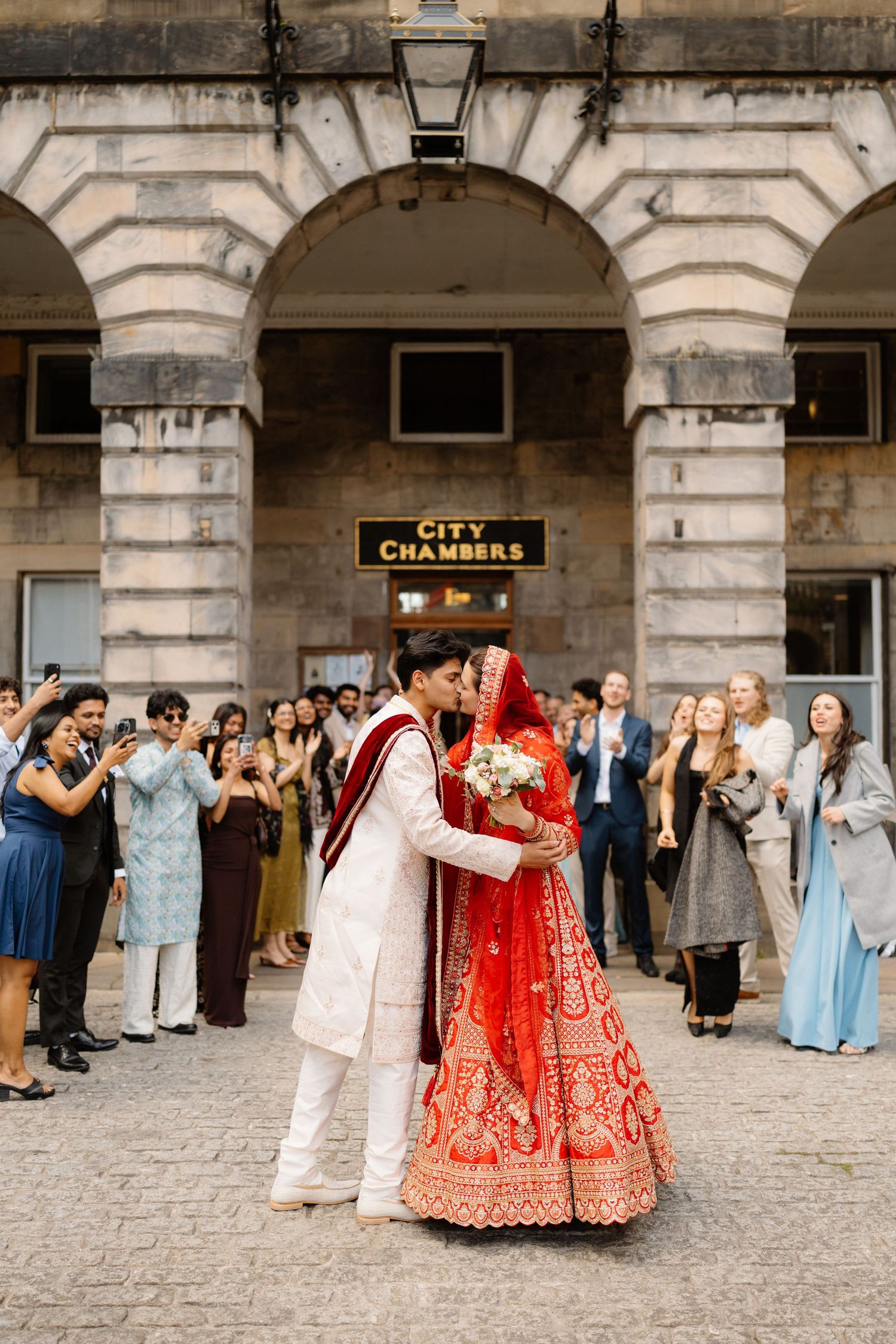 Indian bride in red lehenga and groom in white sherwani share a kiss outside City Chambers, Edinburgh.