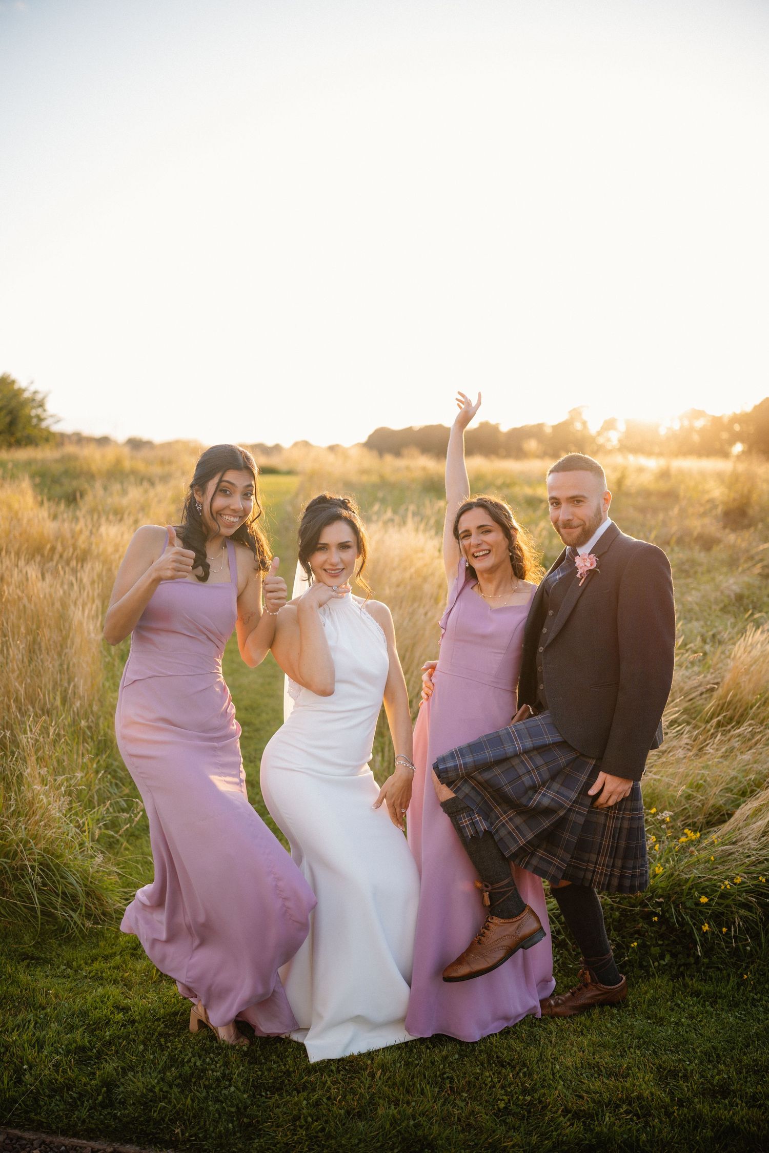 Wedding party sitting and laughing together in a grassy field at sunset.