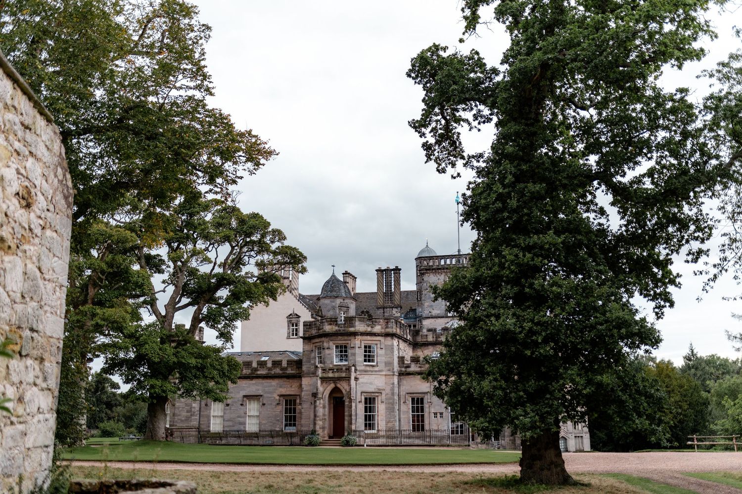 Historic stone mansion with castle-like architecture viewed through mature trees.