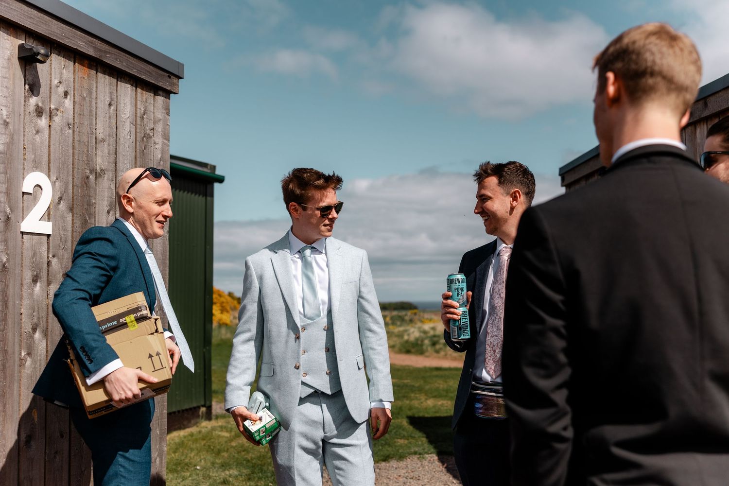 Groomsmen in suits share drinks and laughs outdoors on a sunny day.