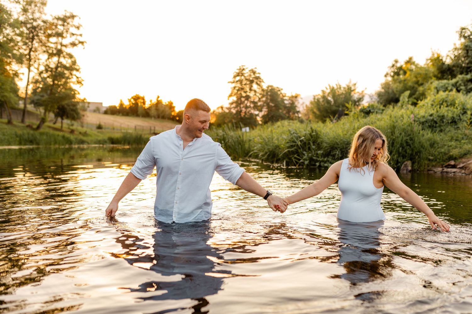 Couple holding hands while wading in sunlit water during sunset.