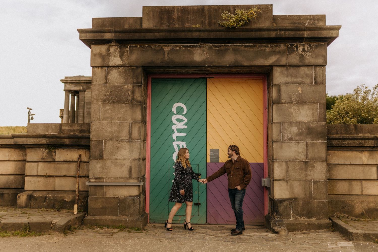 A couple poses playfully in front of colorful doors on a stone building.